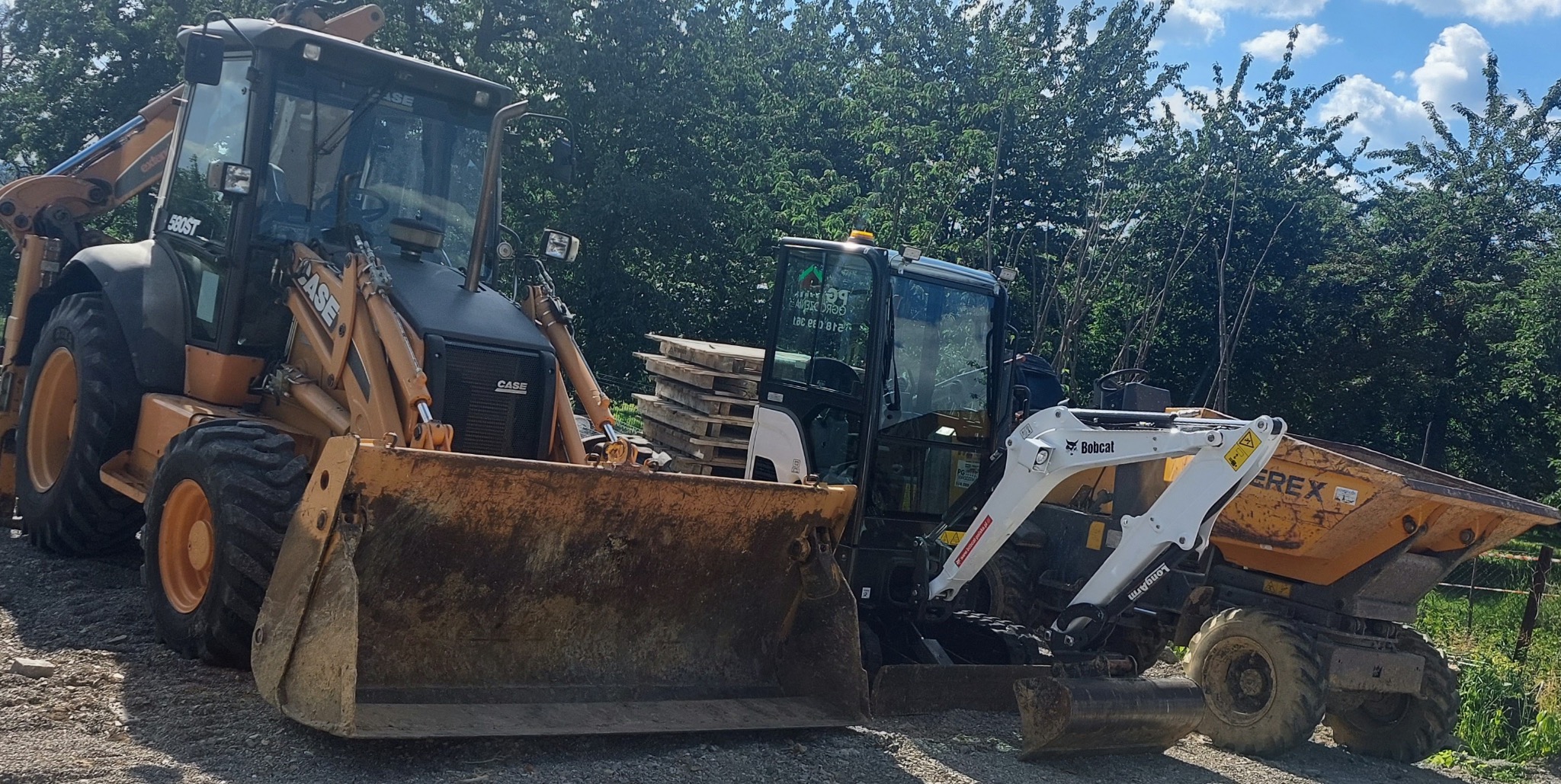 Ciężki sprzęt budowlany: koparko-ładowarka Case 580ST z dużą łyżką, mini koparka Bobcat i wywrotka Terex, stojące obok siebie na żwirowym podłożu, w tle zielone drzewa.