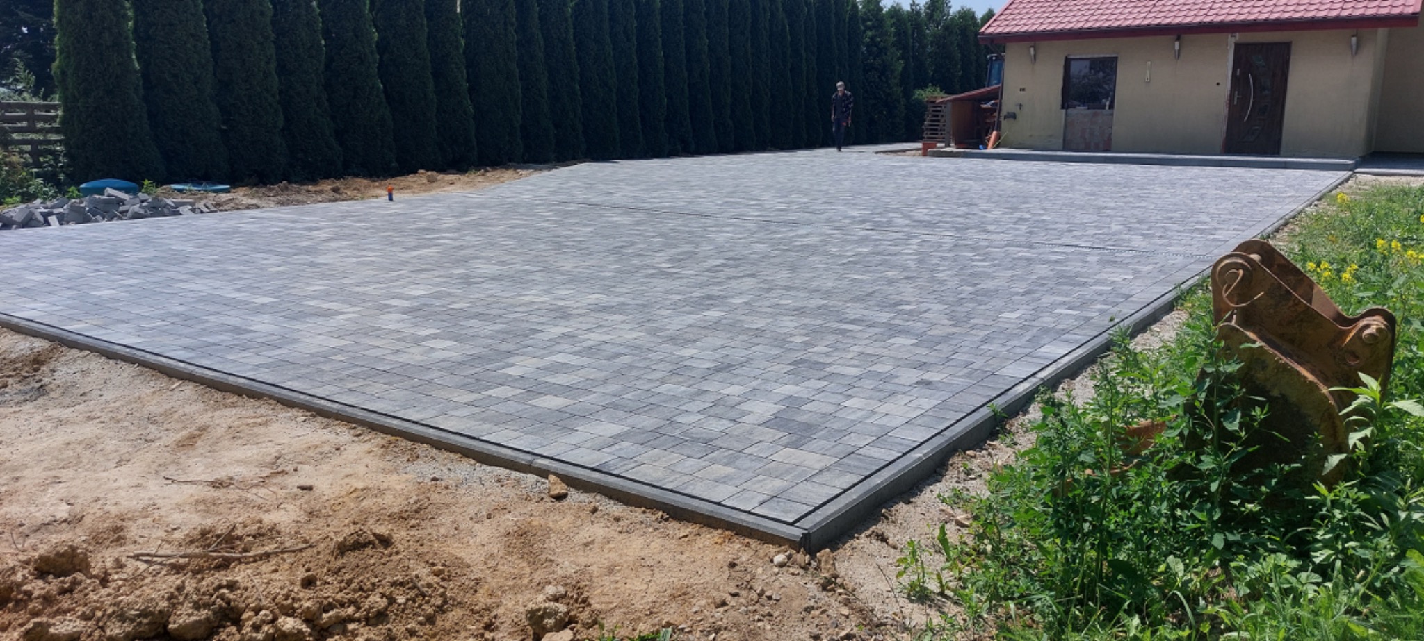 Ukończona nawierzchnia z szarej kostki brukowej ułożonej we wzór, widoczna z perspektywy ziemi, z fragmentem łyżki koparki w prawym dolnym rogu i budynkiem w tle.