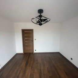 Wesbud - Pusty pokój po odświeżeniu: ciemna drewniana podłoga, białe ściany, nowoczesna czarna lampa sufitowa z białymi elementami, drewniane drzwi z poziomymi panelami.