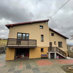 Wesbud - Dwukondygnacyjny dom z beżową elewacją, brązowym dachem i balkonem, widoczny na tle pochmurnego nieba. Otwarty garaż z widocznymi wiadrami farb i osobą w środku, sugerujący prace malarskie.