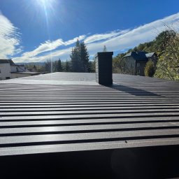 Wesbud - Nowy, brązowy dach z blachy trapezowej z kominem na tle błękitnego nieba z obłokami i zabudowy jednorodzinnej.