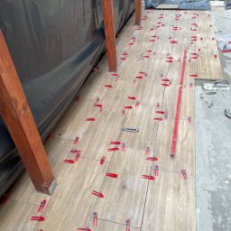 Wesbud - Układanie płytek podłogowych imitujących drewno na tarasie z użyciem czerwonych klipsów poziomujących, widoczny poziomica i nóż do cięcia płytek, czarna folia w tle.