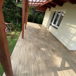 Wesbud - Zadaszony taras z drewnianymi słupami i beżową podłogą imitującą drewno, widok na ogród z huśtawką.