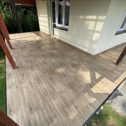 Wesbud - Wyłożony płytkami taras z drewnianymi słupami, widoczne cienie konstrukcji i drzew, jasna elewacja budynku z oknem.