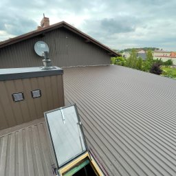 Wesbud - Nowy, brązowy dach z blachy trapezowej z otwartym oknem dachowym i kominem, widok z góry na tle zabudowań i zieleni.