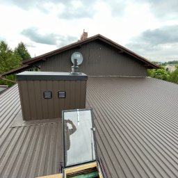 Wesbud - Brązowy dach z blachy trapezowej z zamontowanym wyłazem dachowym, kominem wentylacyjnym i anteną satelitarną, widok z góry na tle pochmurnego nieba.