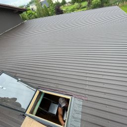 Wesbud - Montaż okna dachowego na nowym, szarym dachu z blachy trapezowej; widoczny fragment drabiny i pracownik w czapce z daszkiem.