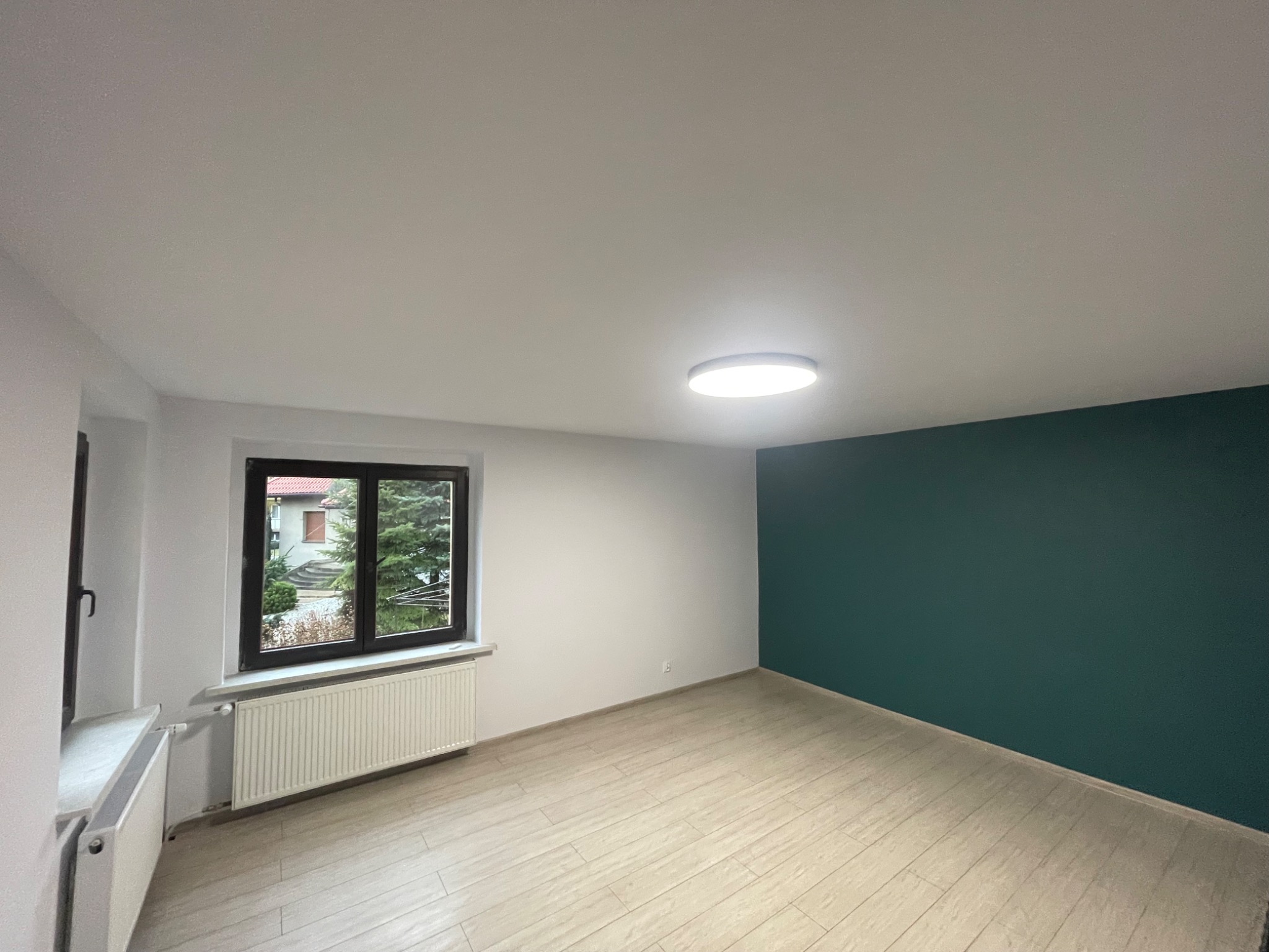 Pomalowane na biało i zielono puste pomieszczenie z grzejnikiem pod oknem z widokiem na ogród i drewnianą podłogą.