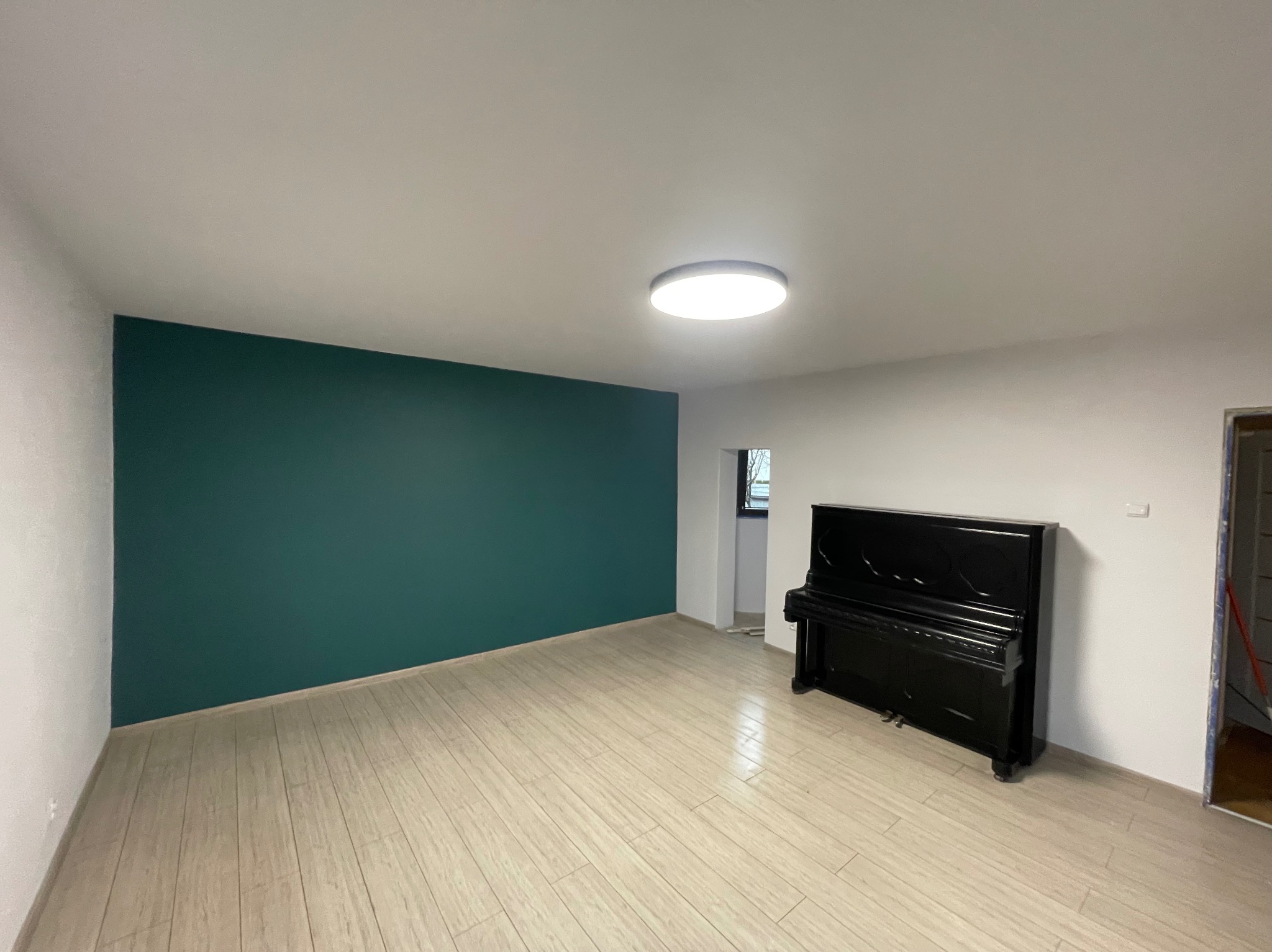 Pokój po remoncie z pomalowaną na zielono ścianą, jasną podłogą imitującą drewno i czarnym pianinem.