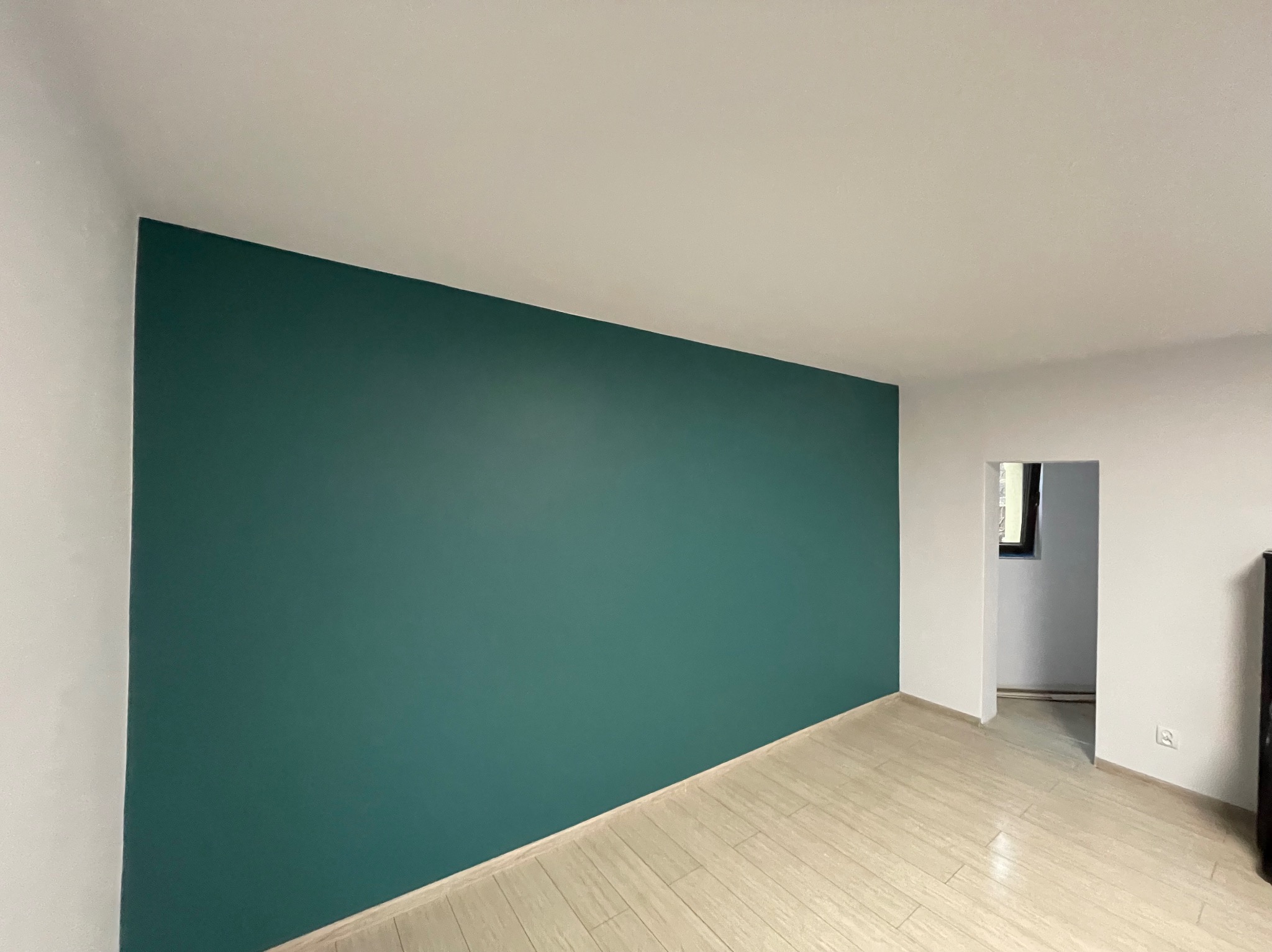 Pomieszczenie po malowaniu: ściana w odcieniu butelkowej zieleni kontrastuje z bielą sufitu i ścian, jasne panele podłogowe, widoczne przejście do kolejnego pomieszczenia.