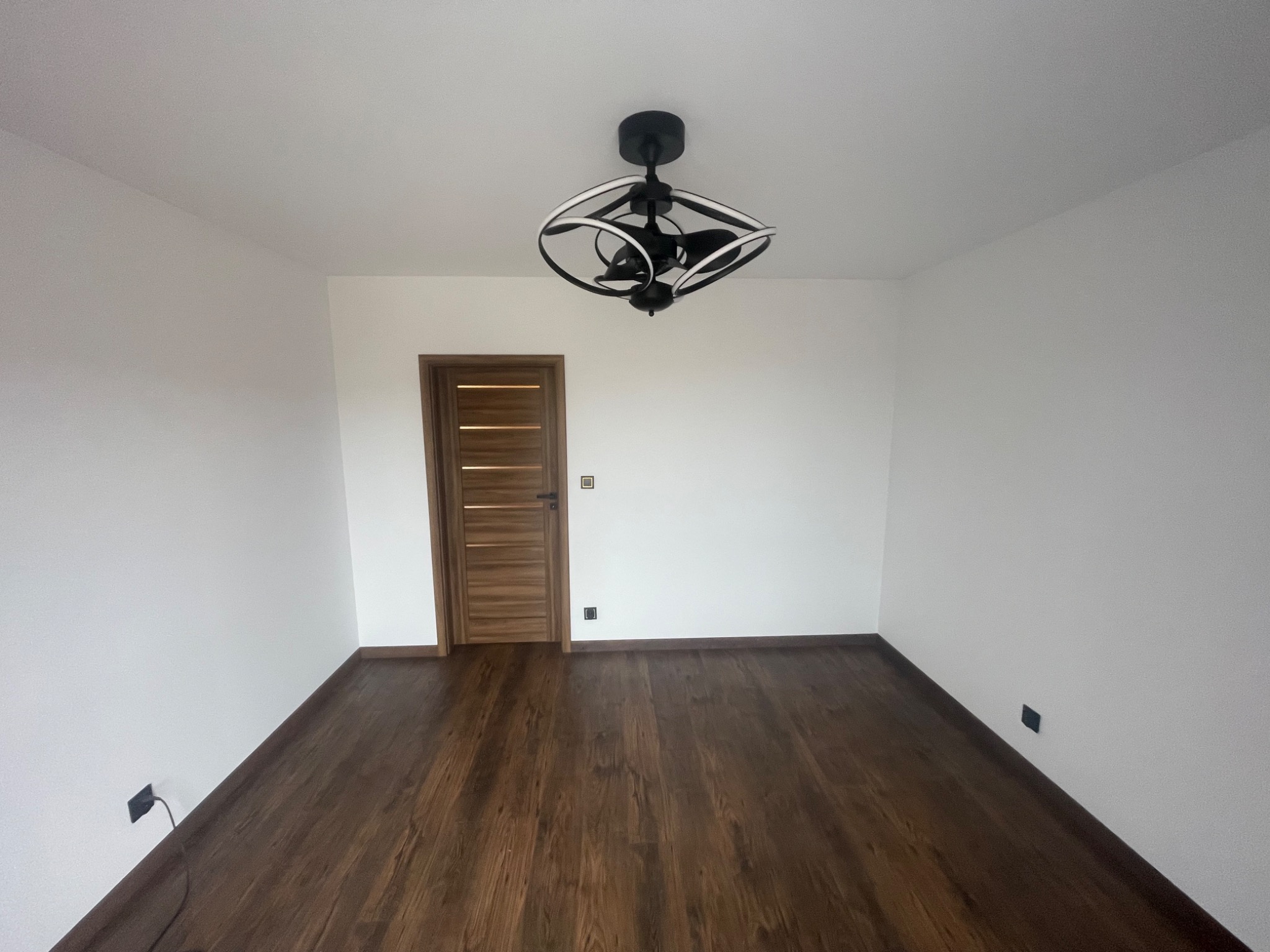 Pusty pokój po odświeżeniu: ciemna drewniana podłoga, białe ściany, nowoczesna czarna lampa sufitowa z białymi elementami, drewniane drzwi z poziomymi panelami.