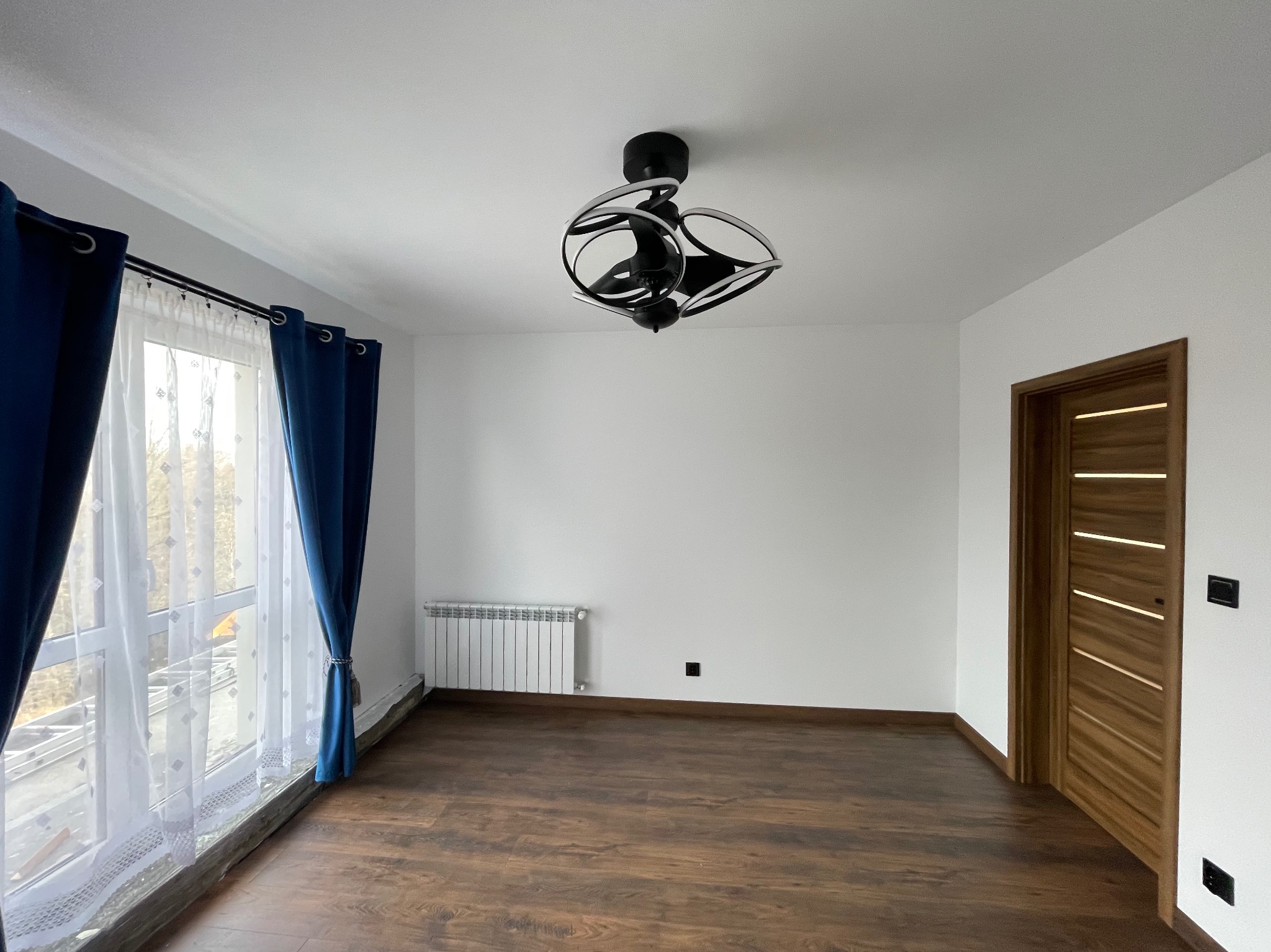 Pusty pokój po remoncie z ciemną podłogą, białymi ścianami, nowoczesną lampą sufitową, grzejnikiem, drewnianymi drzwiami i niebieskimi zasłonami przy oknie.