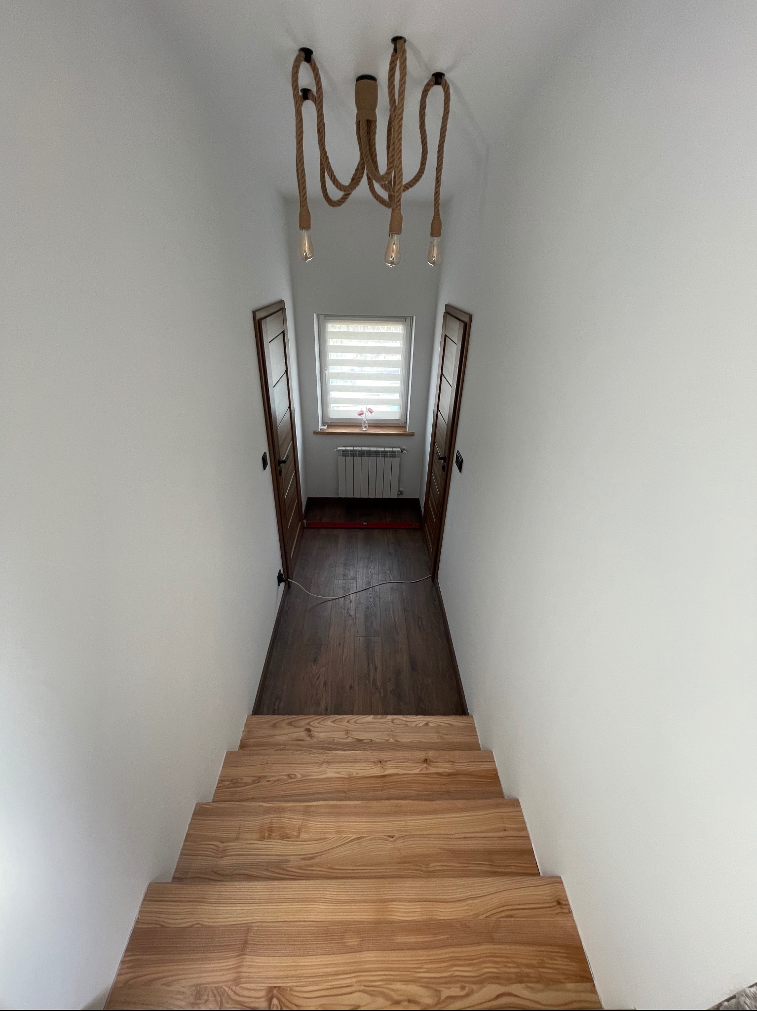 Schody z jasnego drewna prowadzące do korytarza z ciemną podłogą, białe ściany, okno z żaluzjami i lampa w stylu marynistycznym.