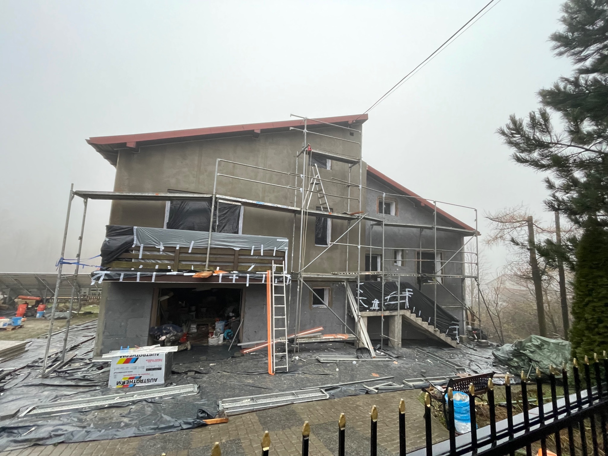 Dom jednorodzinny w trakcie ocieplania, otoczony rusztowaniem, z widocznymi paczkami styropianu Austrotherm, zabezpieczony folią, w tle zalesiony teren w mglisty dzień.