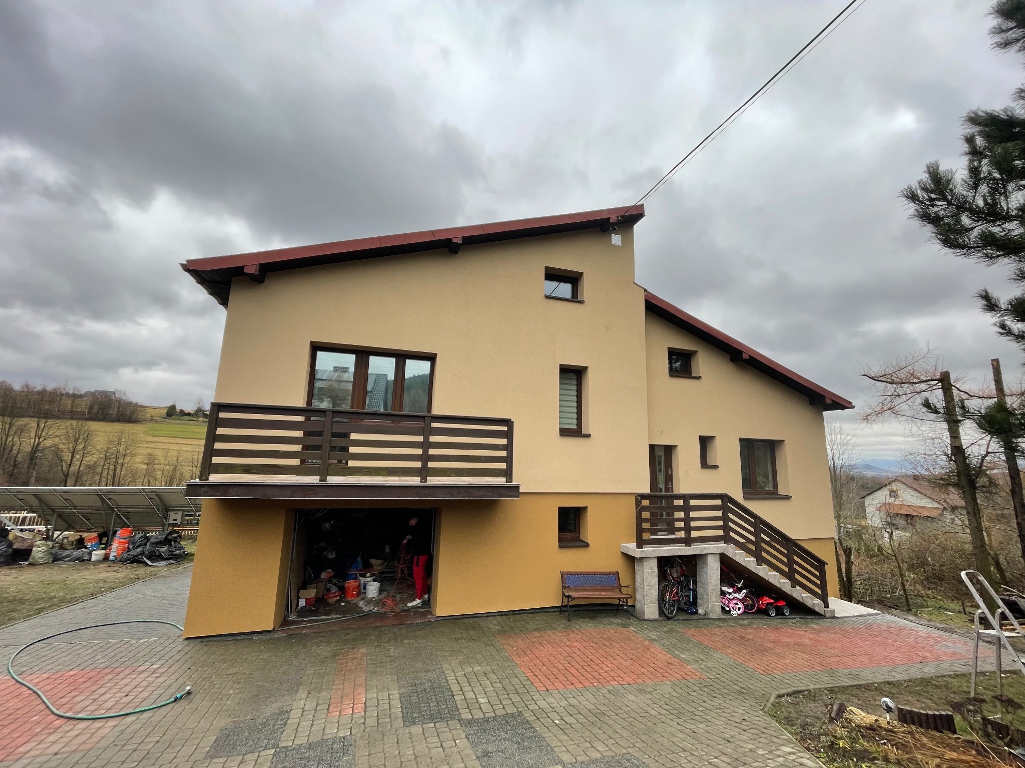 Dwukondygnacyjny dom z beżową elewacją, brązowym dachem i balkonem, widoczny na tle pochmurnego nieba. Otwarty garaż z widocznymi wiadrami farb i osobą w środku, sugerujący prace malarskie.