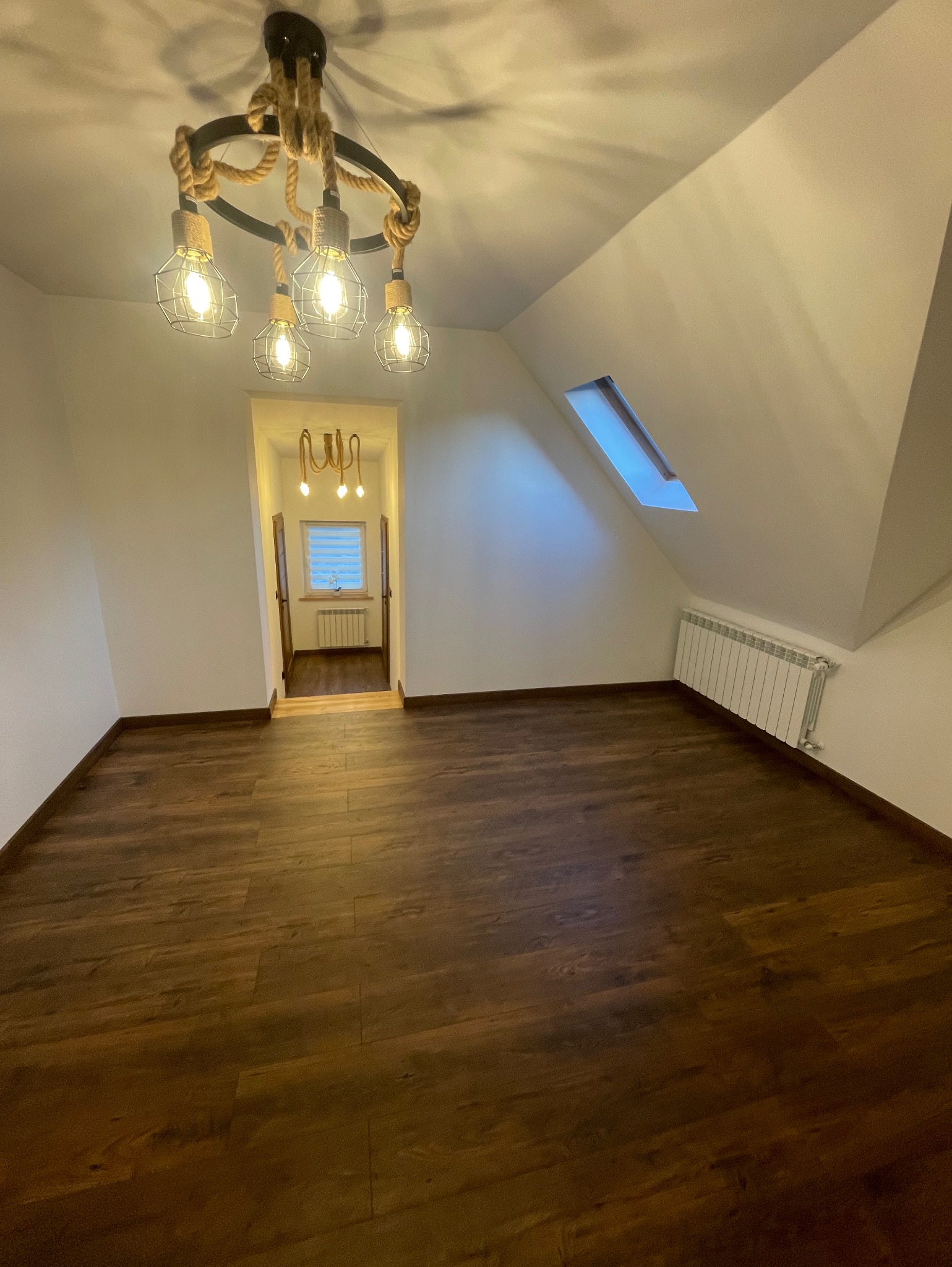 Puste poddasze z drewnianą podłogą, widok na sąsiednie pomieszczenie przez otwarte drzwi, okno dachowe i dekoracyjna lampa wisząca ze sznura.