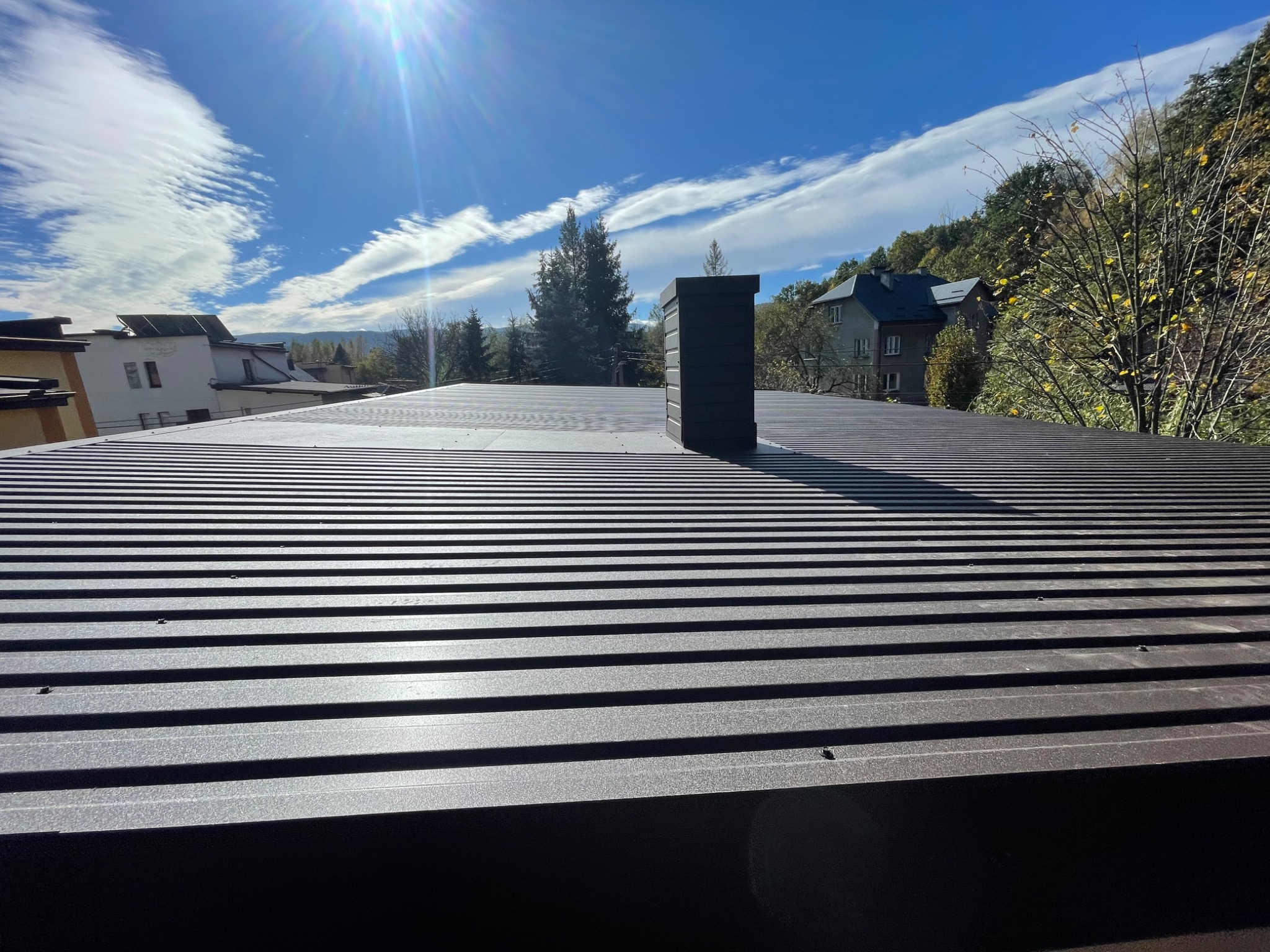 Nowy, brązowy dach z blachy trapezowej z kominem na tle błękitnego nieba z obłokami i zabudowy jednorodzinnej.