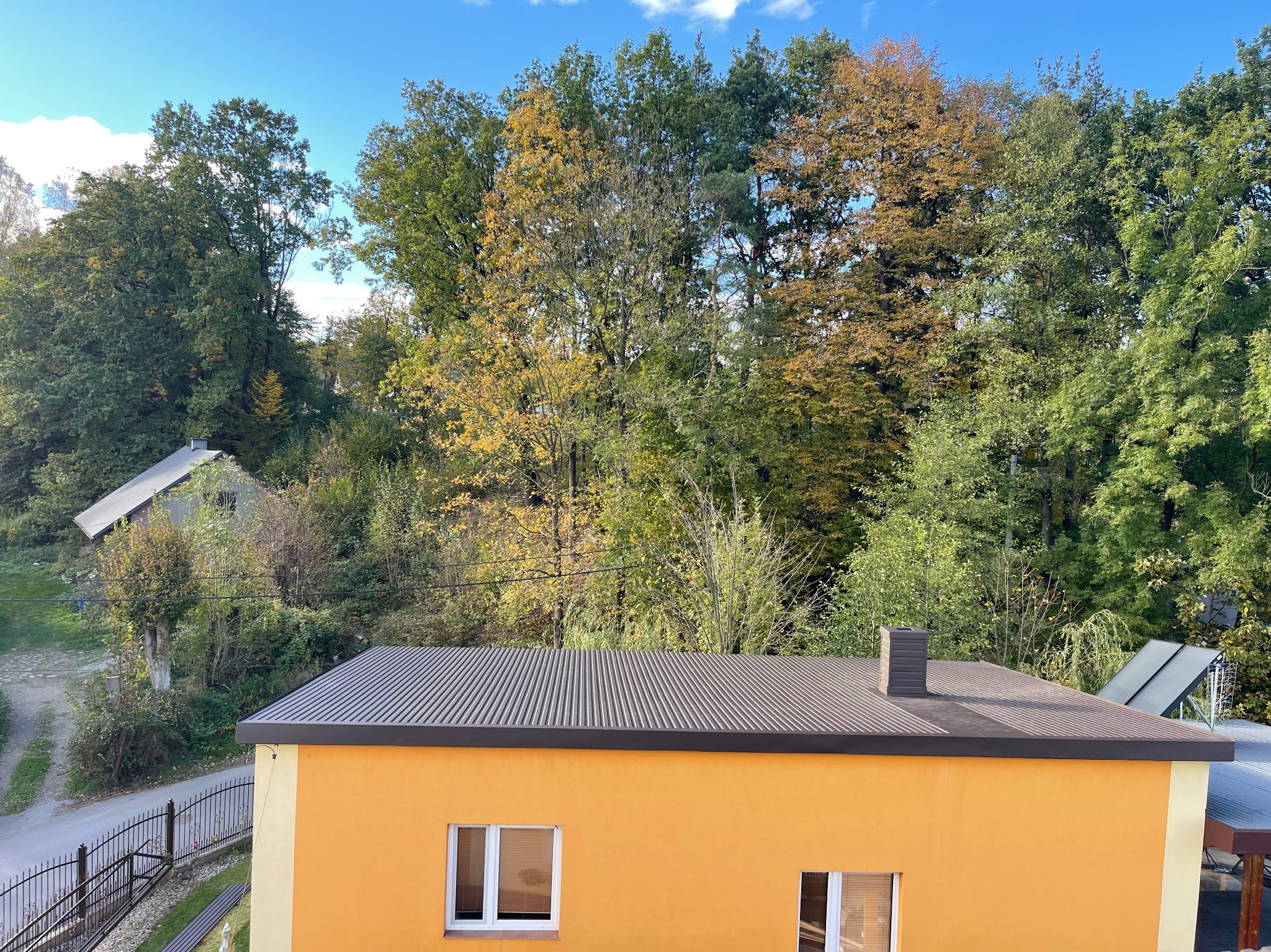 Pomarańczowy dom z nowym, ciemnoszarym dachem na tle jesiennego lasu i błękitnego nieba, widok z góry.