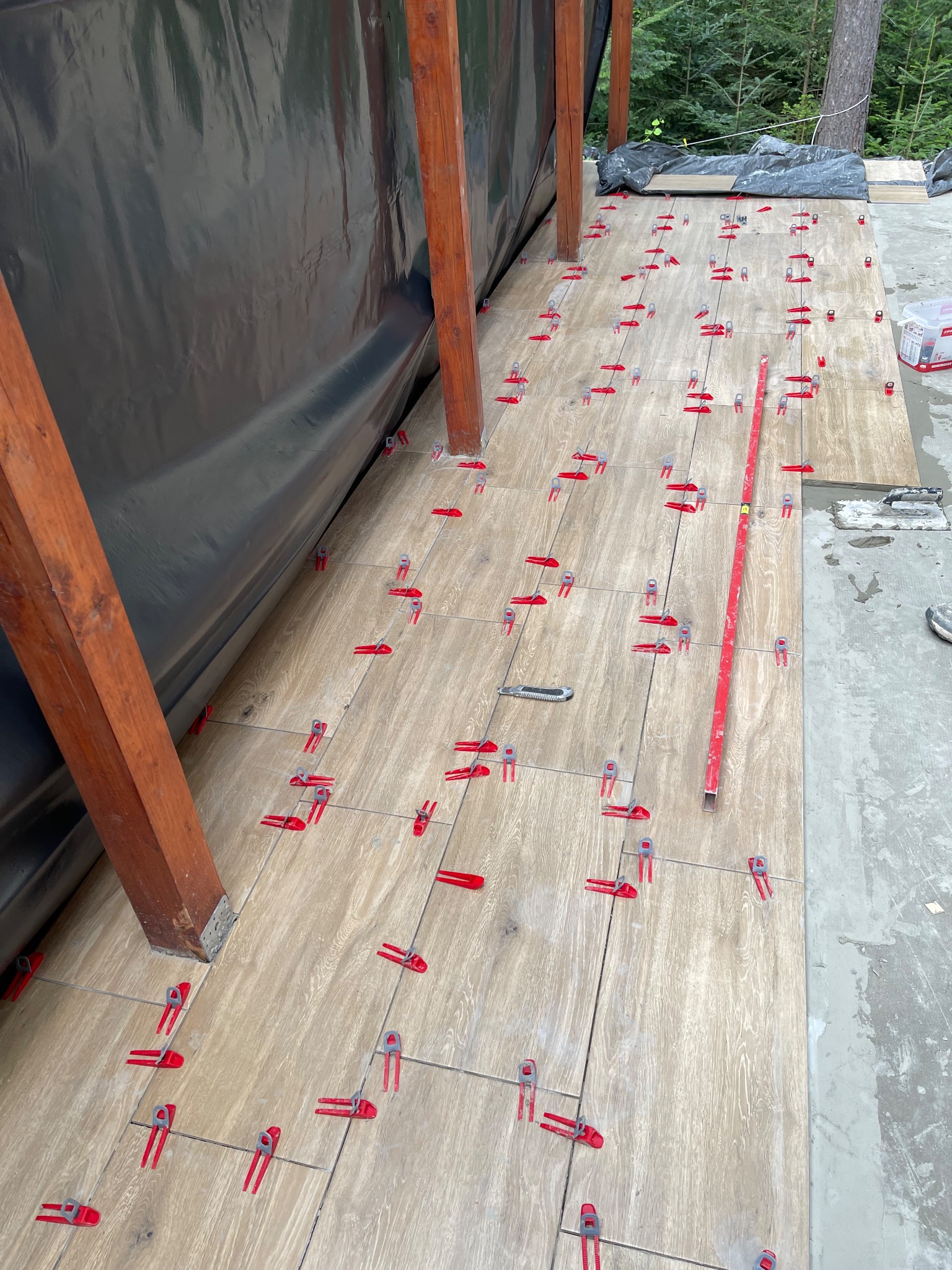 Układanie płytek podłogowych imitujących drewno na tarasie z użyciem czerwonych klipsów poziomujących, widoczny poziomica i nóż do cięcia płytek, czarna folia w tle.
