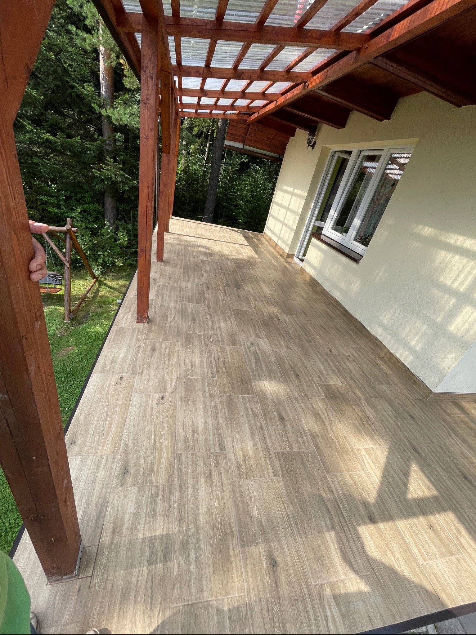 Zadaszony taras z drewnianymi słupami i beżową podłogą imitującą drewno, widok na ogród z huśtawką.