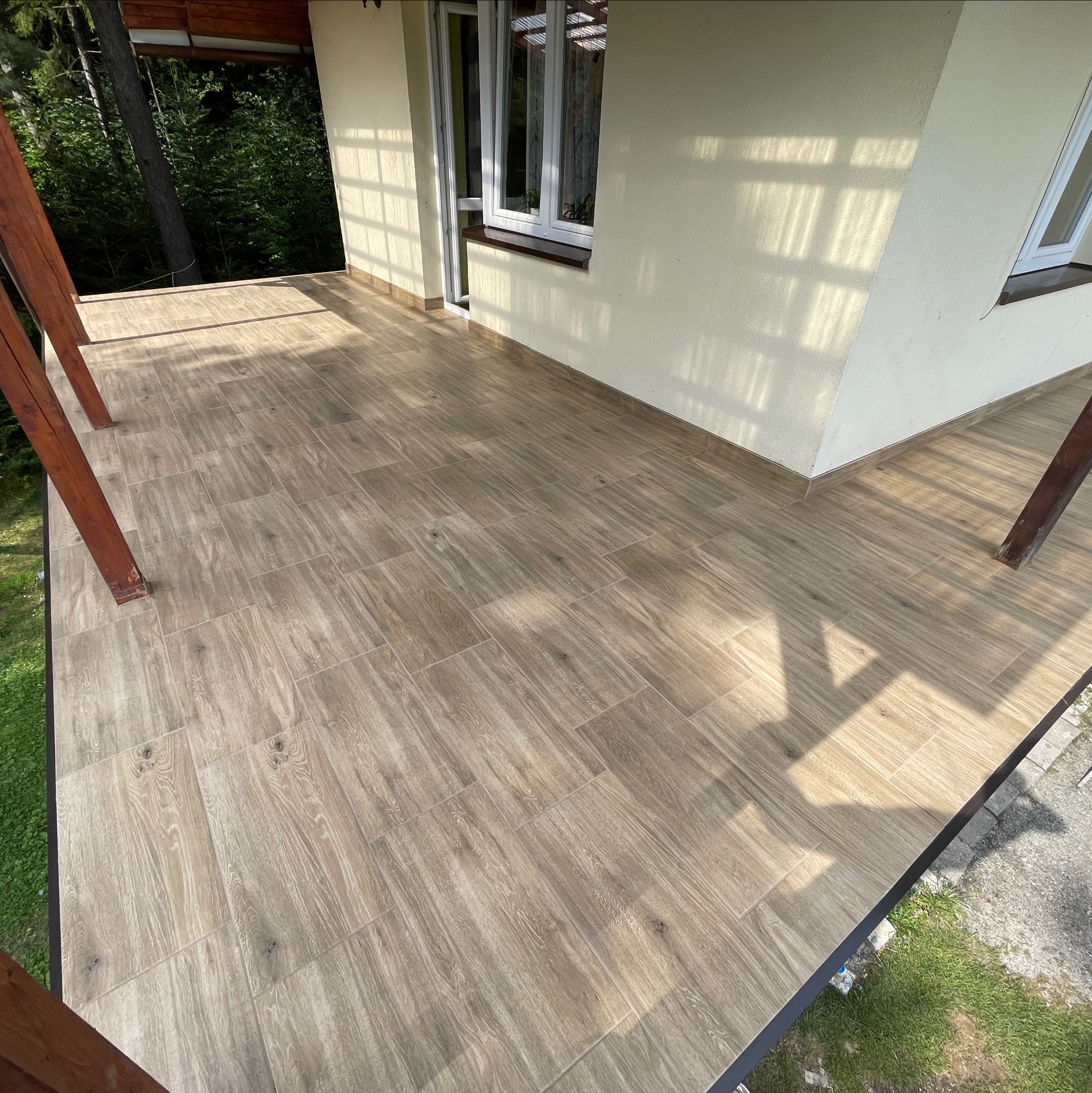 Wyłożony płytkami taras z drewnianymi słupami, widoczne cienie konstrukcji i drzew, jasna elewacja budynku z oknem.