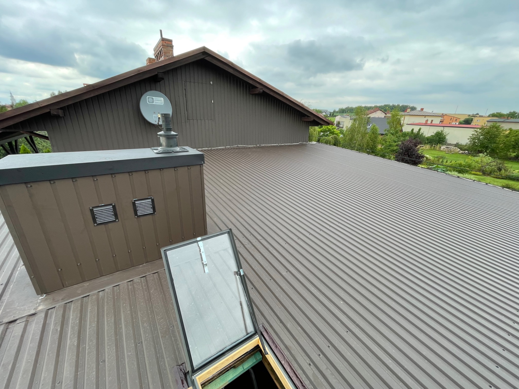 Nowy, brązowy dach z blachy trapezowej z otwartym oknem dachowym i kominem, widok z góry na tle zabudowań i zieleni.