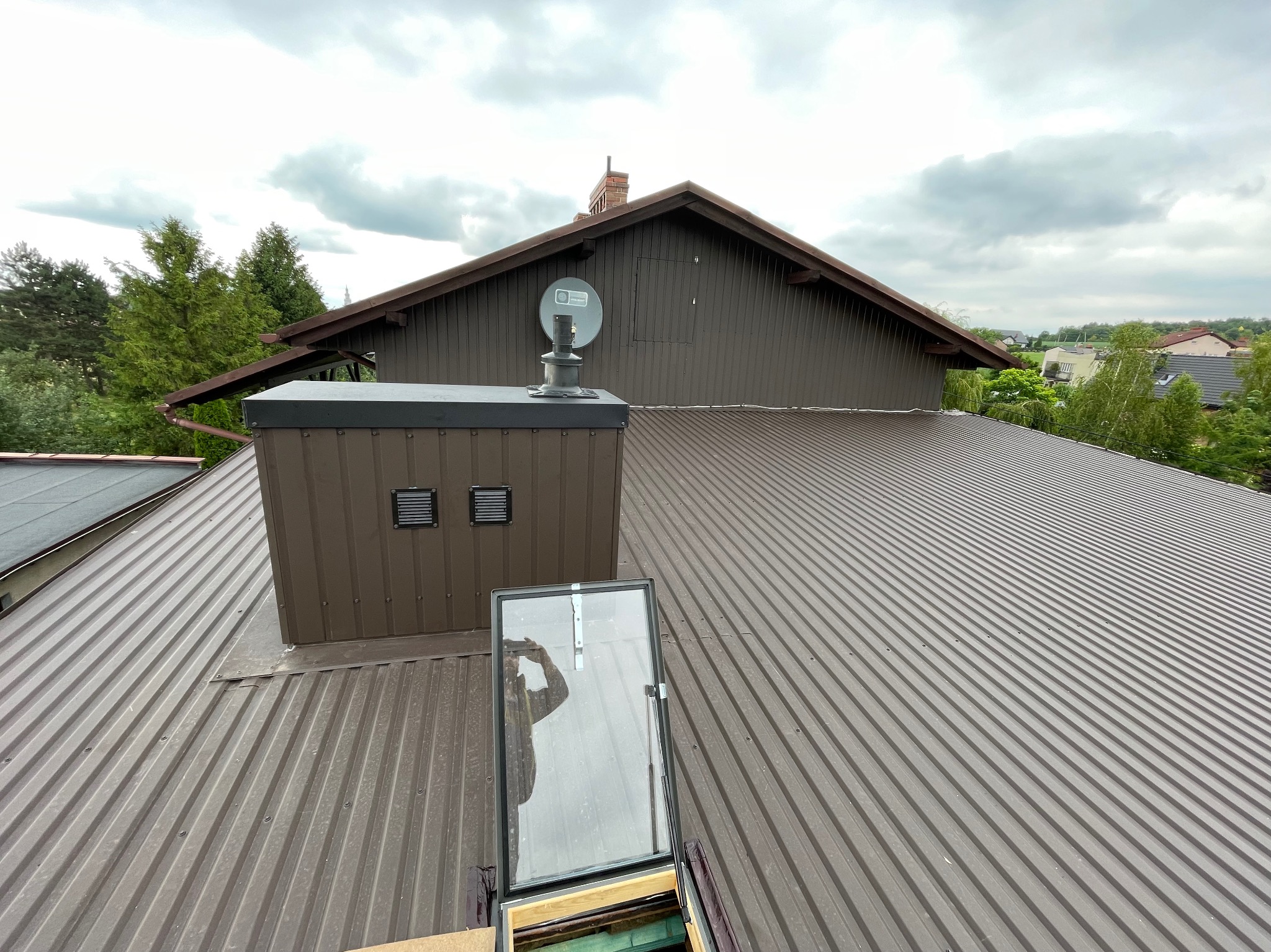 Brązowy dach z blachy trapezowej z zamontowanym wyłazem dachowym, kominem wentylacyjnym i anteną satelitarną, widok z góry na tle pochmurnego nieba.