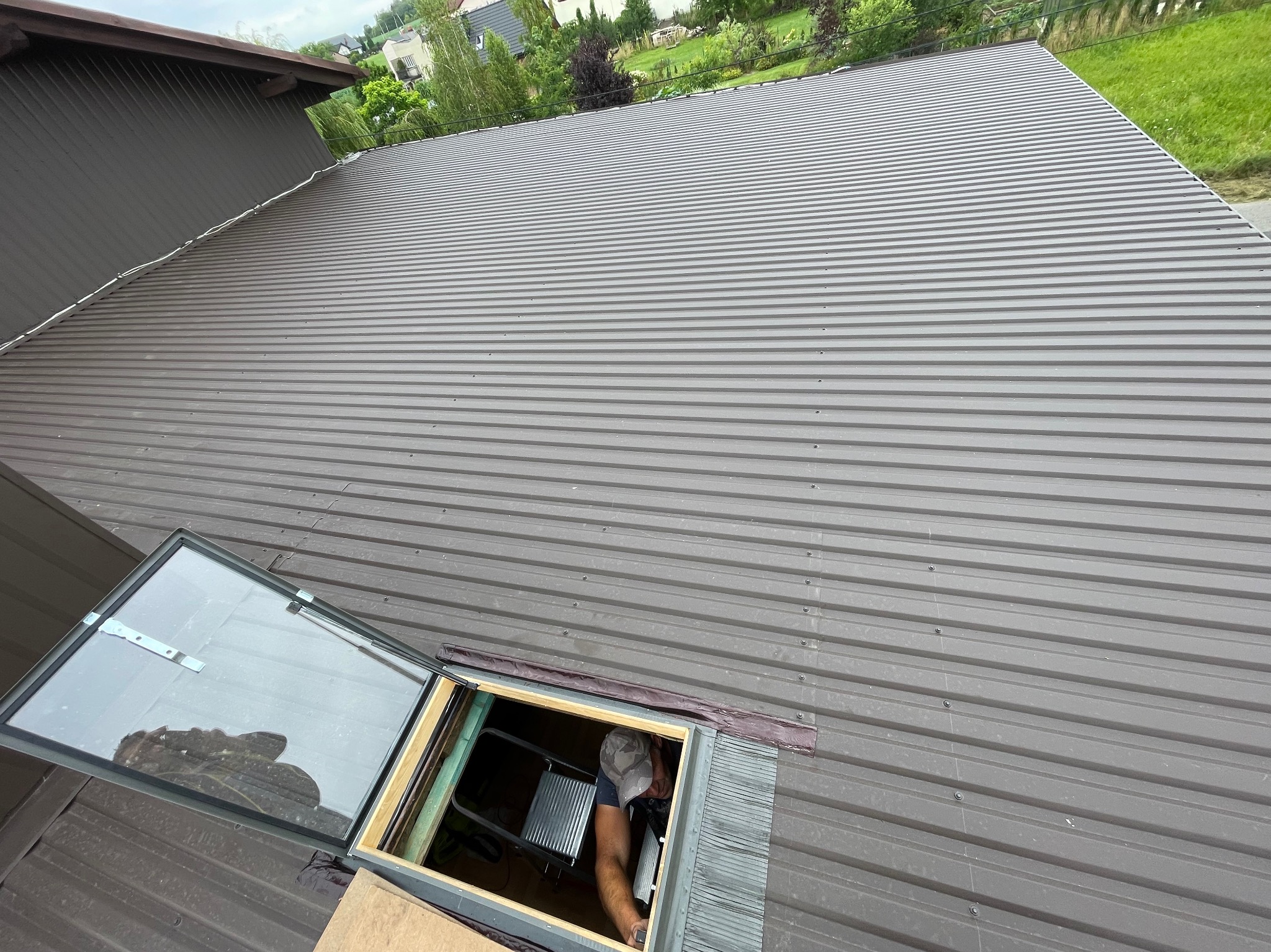 Montaż okna dachowego na nowym, szarym dachu z blachy trapezowej; widoczny fragment drabiny i pracownik w czapce z daszkiem.