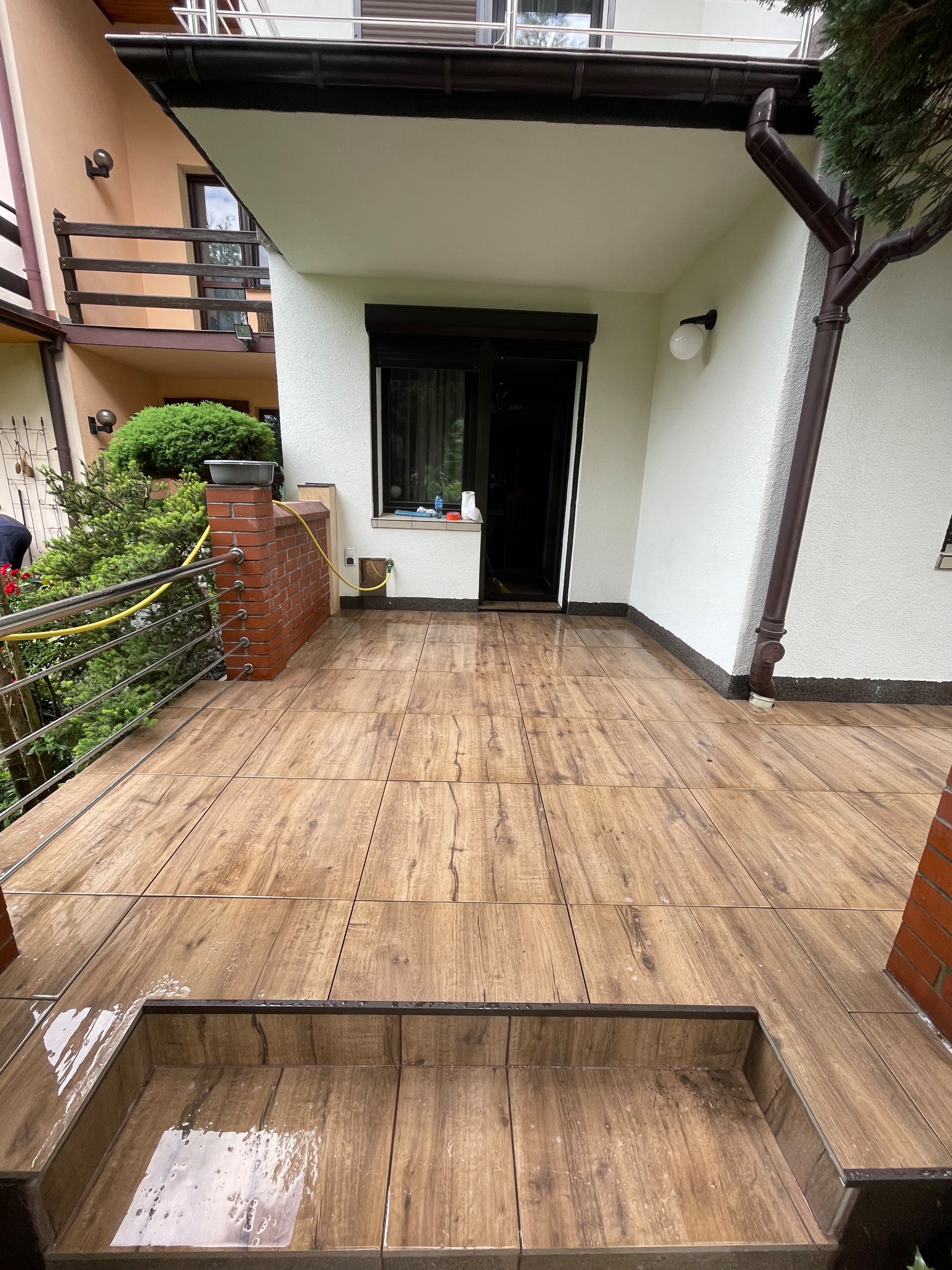 Wyłożony drewnopodobnymi płytkami taras z dwoma stopniami, częściowo mokry, z widoczną elewacją budynku i rynną.