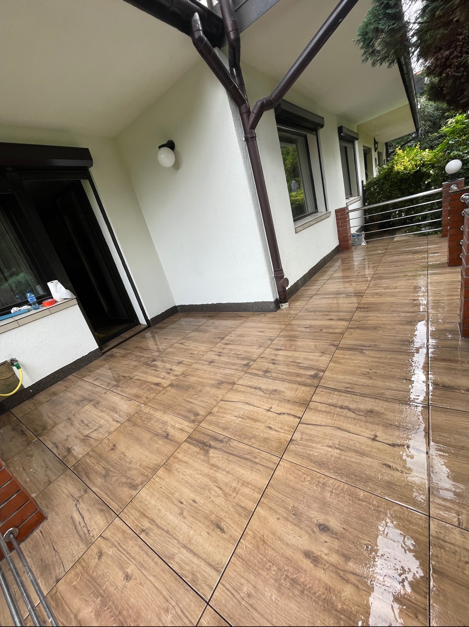 Umyta, brązowa, drewnopodobna terakota na balkonie domu jednorodzinnego, widoczne mokre ślady po myciu, na ścianie biały tynk i brązowe rynny.