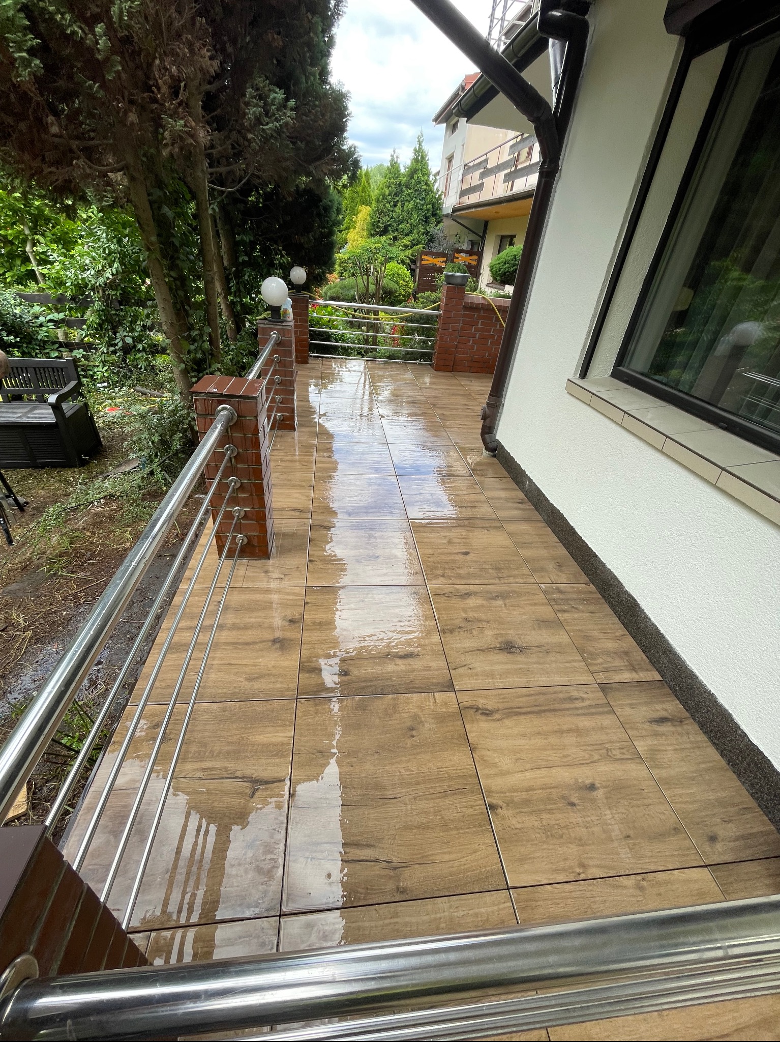 Mokry taras z drewnopodobnych płytek, ogrodzony metalową balustradą z ceglanymi słupkami, przylegający do białej ściany budynku.