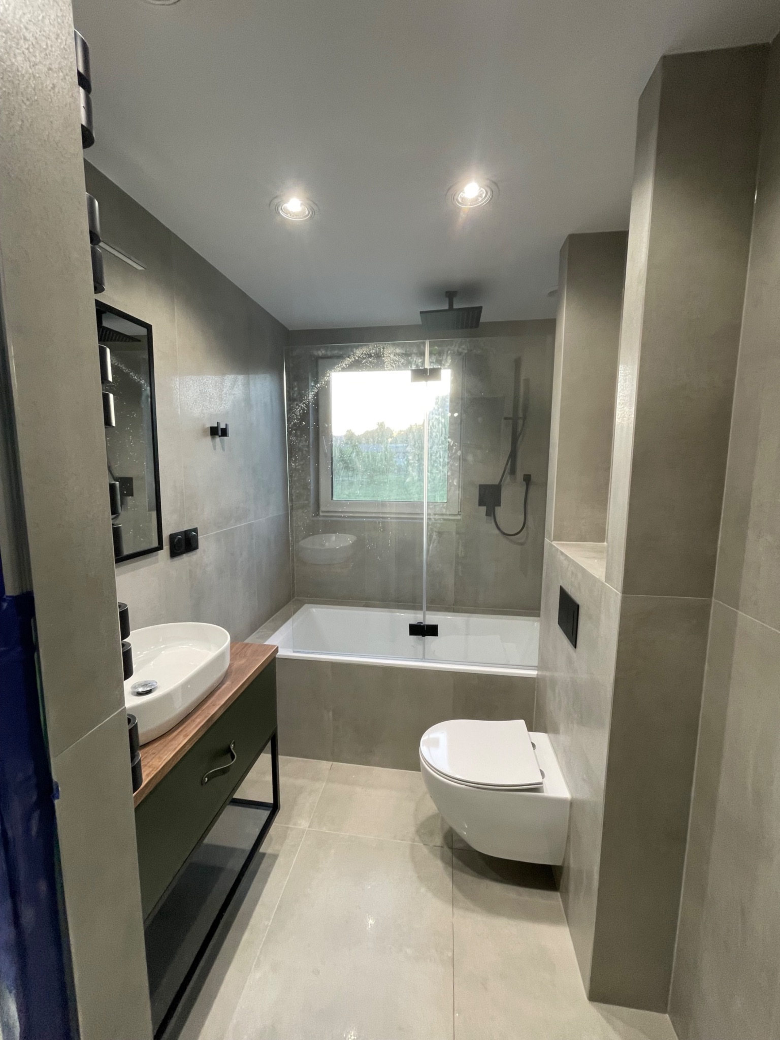 Nowoczesna łazienka w odcieniach szarości z białą armaturą i drewnianym blatem pod umywalką; widoczna wanna za szybą, toaleta podwieszana i minimalistyczne dodatki.