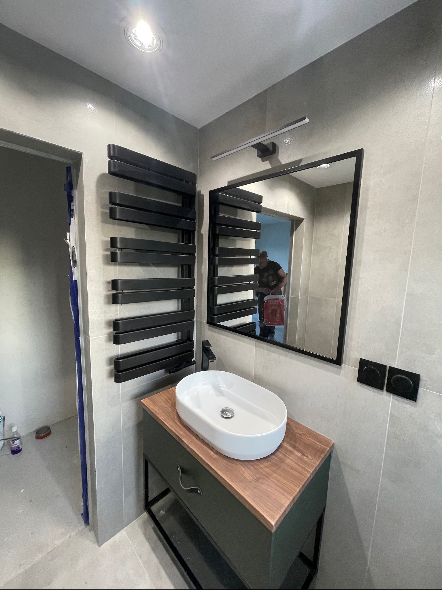 Nowoczesna łazienka z szarymi płytkami, wiszącym grzejnikiem drabinkowym w czarnym kolorze, umywalką nablatową na szafce z drewnianym blatem i czarną armaturą, oraz lustrem w czarnej ramie,...