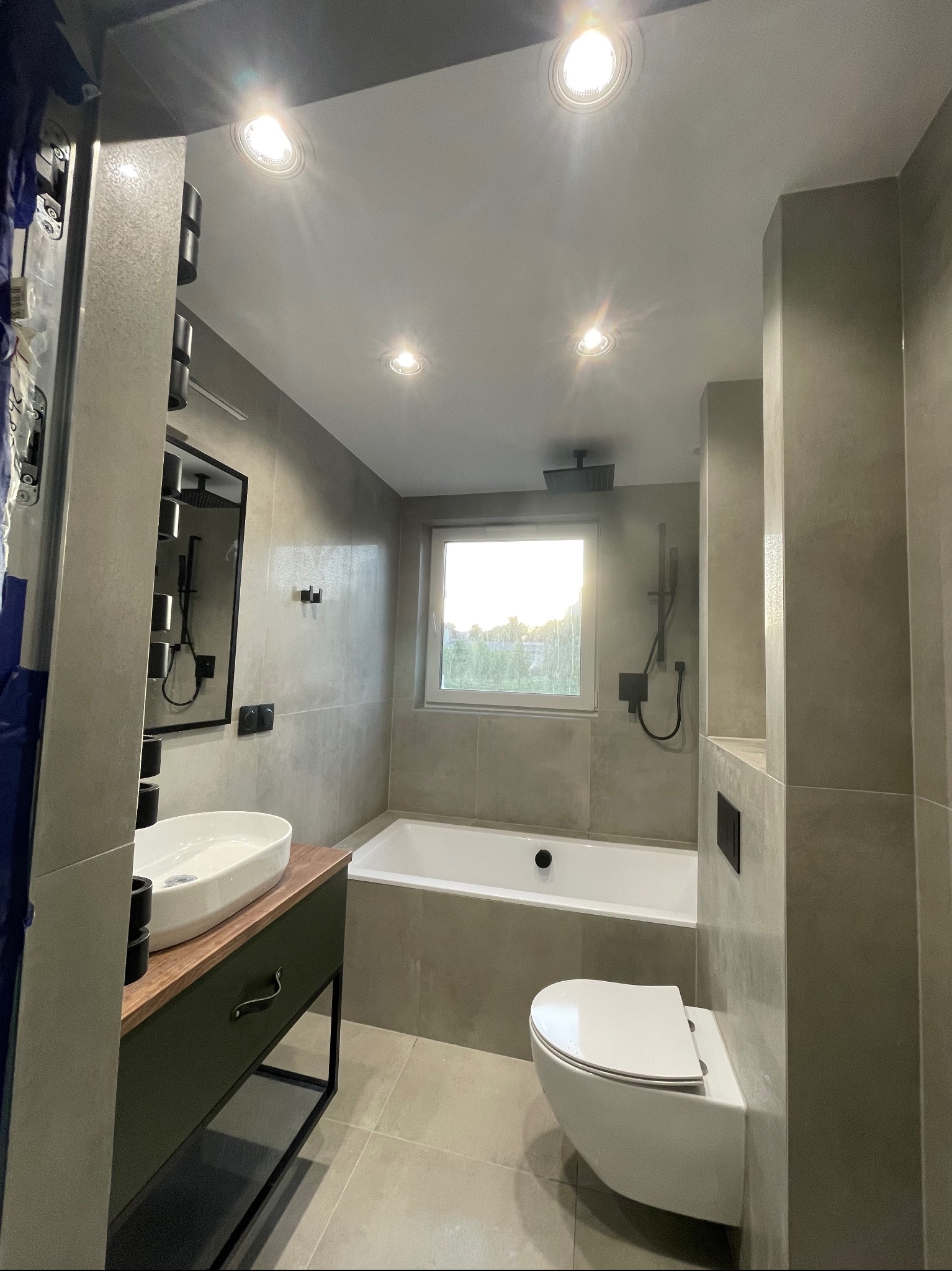 Nowoczesna łazienka w szarościach z wanną, podwieszaną toaletą i umywalką na szafce z drewnianym blatem, oświetlona punktowymi lampami sufitowymi.