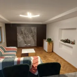 Salon po metamorfozie z parkietem ułożonym w jodełkę, szarą ścianą akcentową z imitacją marmuru, nowoczesnym oświetleniem sufitowym i wnęką z dekoracjami świątecznymi.