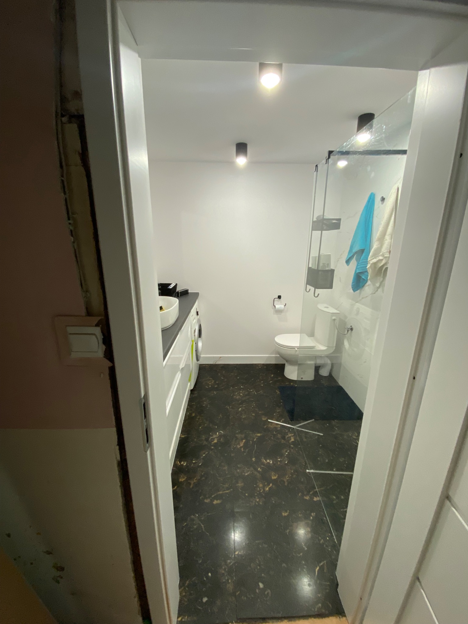 Widok na wyremontowaną łazienkę z czarną, marmurkową podłogą, białą armaturą i szklaną kabiną prysznicową, widoczne niedokończone wykończenie ościeżnicy drzwi.