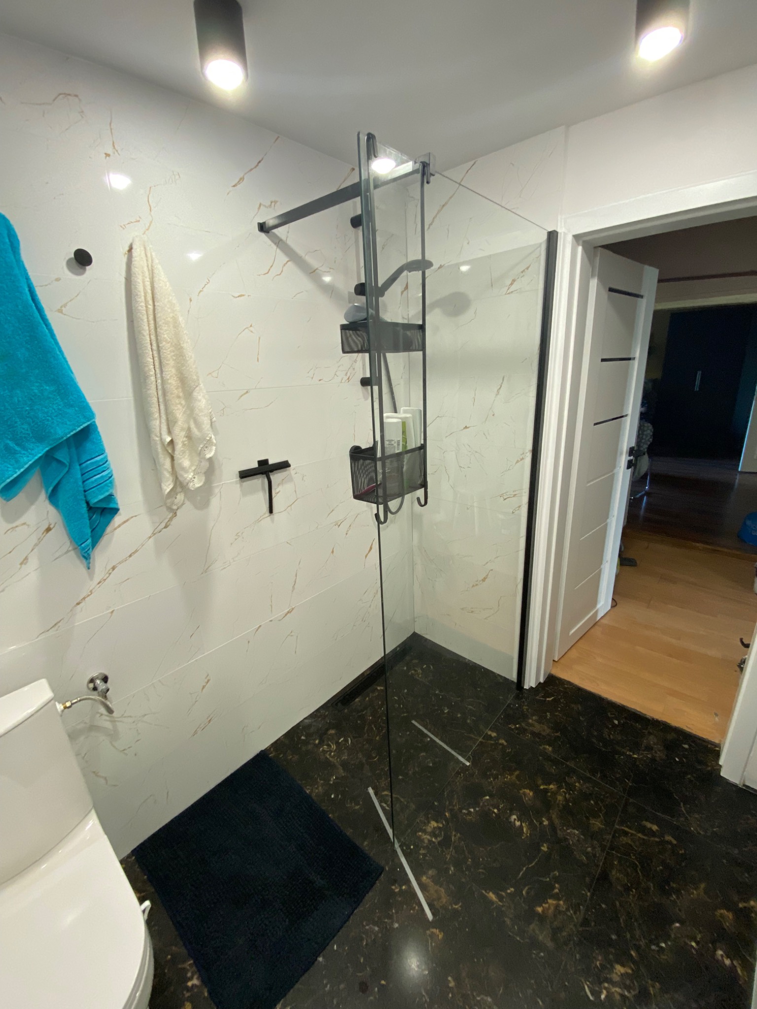 Nowoczesna kabina prysznicowa z czarnymi elementami wykończeniowymi, marmurowe płytki na ścianach i podłodze, widoczny fragment toalety i drzwi do innego pomieszczenia.