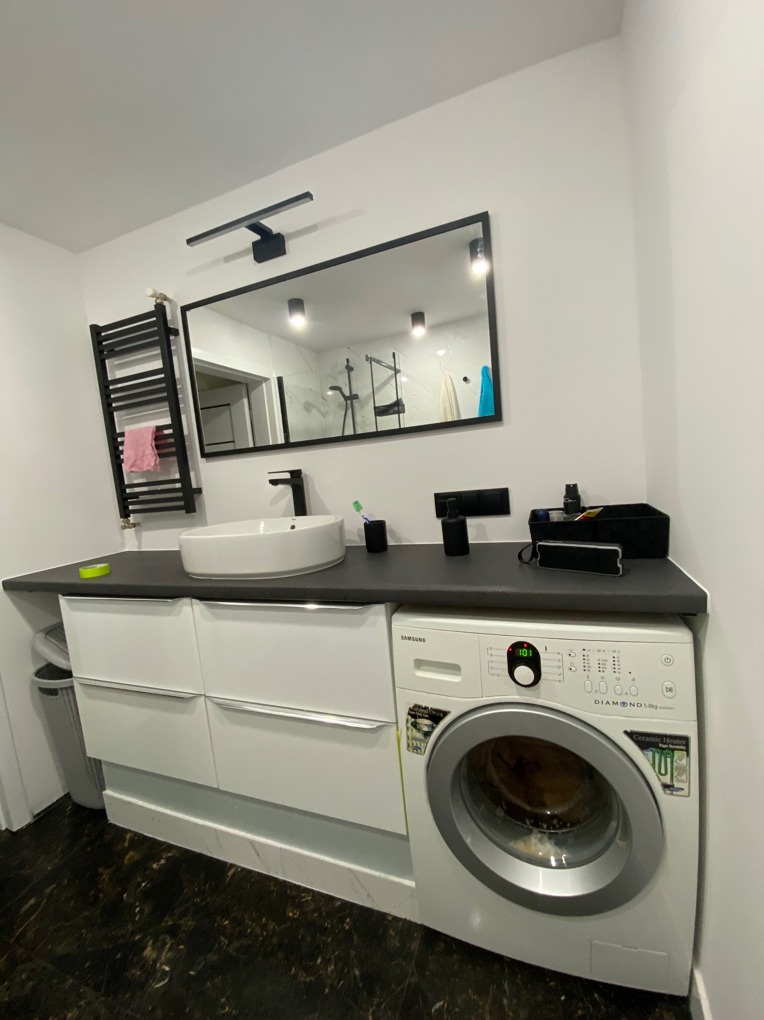 Nowoczesna łazienka z czarnymi akcentami: umywalka nablatowa, lustro w czarnej ramie, grzejnik drabinkowy i pralka Samsung, marmurowa podłoga.