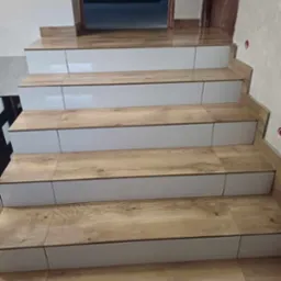 Schody wykończone płytkami imitującymi drewno z białymi panelami bocznymi, prowadzące do drzwi z ościeżnicą w kolorze drewna.