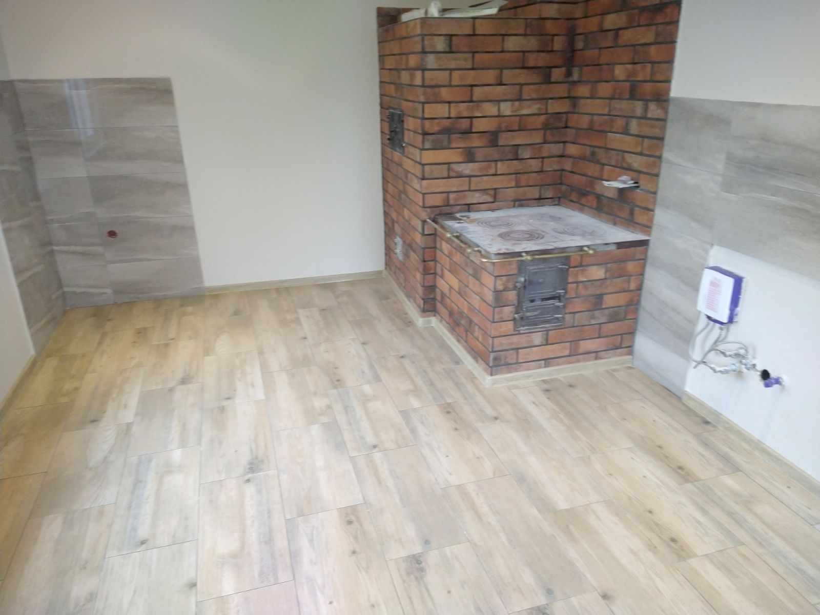 Wnętrze po remoncie z nową podłogą imitującą drewno, fragmentem ściany wyłożonej szarą płytką oraz ceglanym piecem kuchennym z żeliwną płytą grzewczą.
