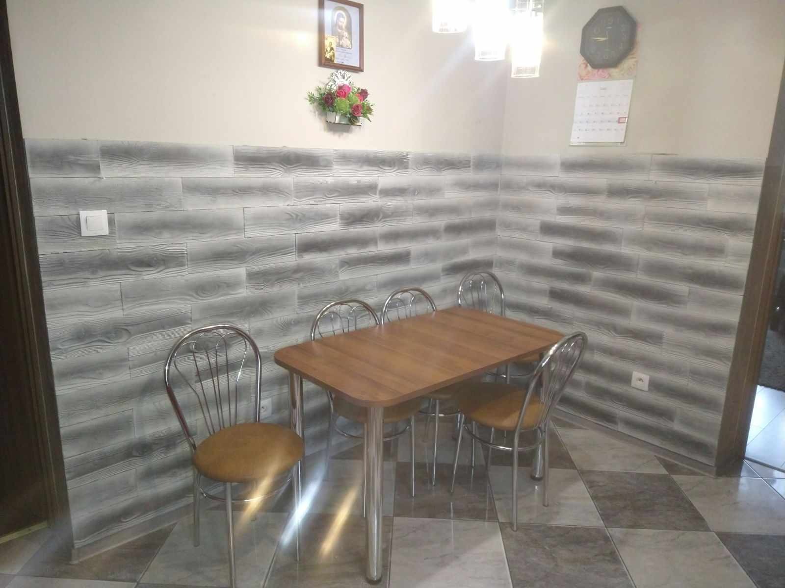 Jadalnia z drewnianym stołem i czterema krzesłami na tle ściany wyłożonej panelami imitującymi drewno, nad stołem wisi obrazek i dekoracja kwiatowa.