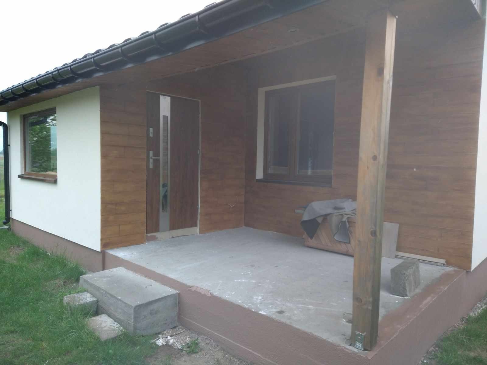 Wykończona elewacja domu z drewnianą okładziną i białym tynkiem, widoczny betonowy taras wejściowy z dwoma stopniami i drewniany słup podtrzymujący zadaszenie.