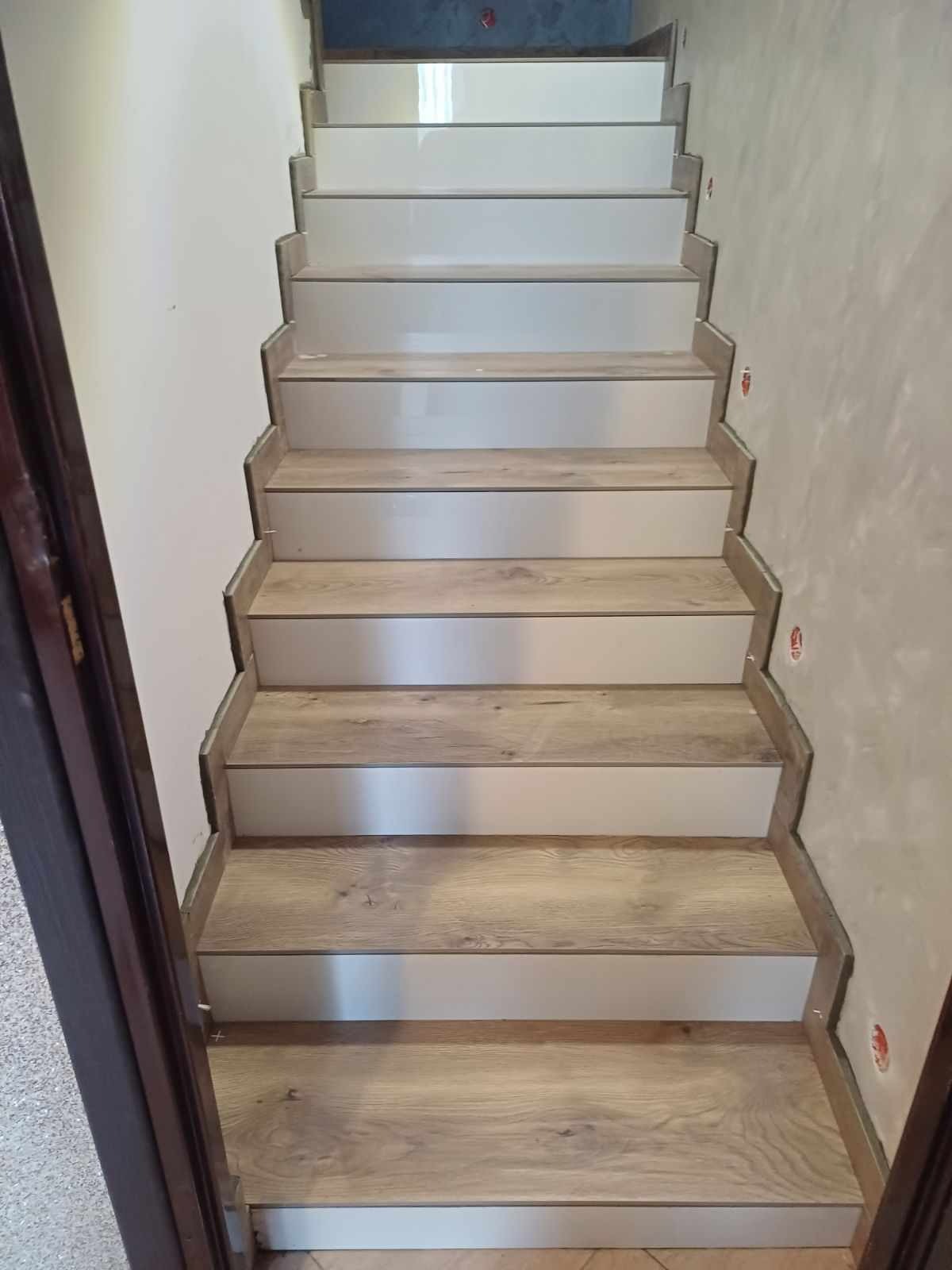 Wykończone schody z drewnianymi stopniami i białymi podstopnicami, widok z dołu, ściana w trakcie malowania, widoczne ślady po gipsowaniu.
