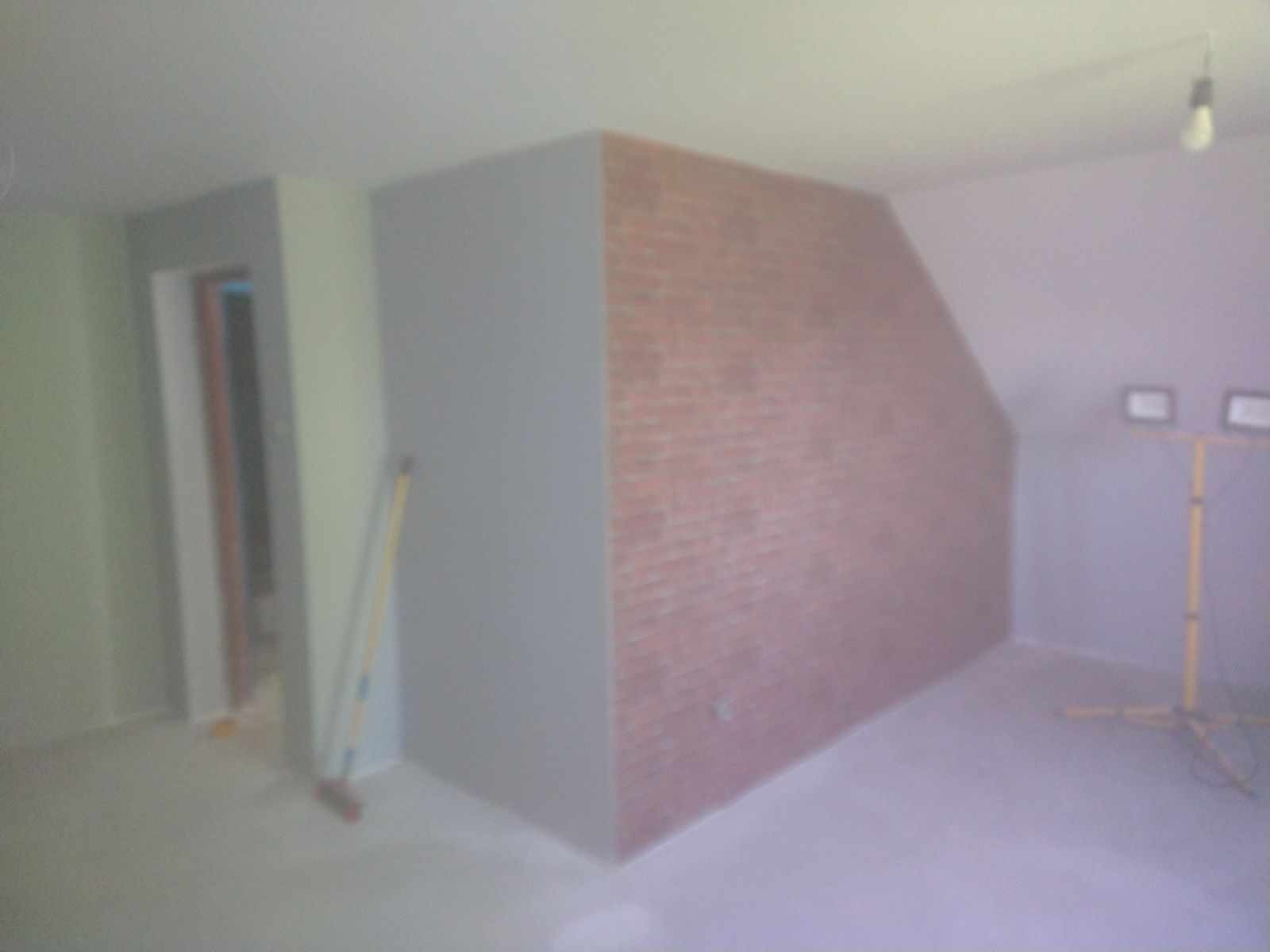 Wnętrze w trakcie prac wykończeniowych: ściana z cegieł dekoracyjnych, szara ściana, poziomica oparta o ścianę, lampa budowlana na statywie, surowa podłoga.