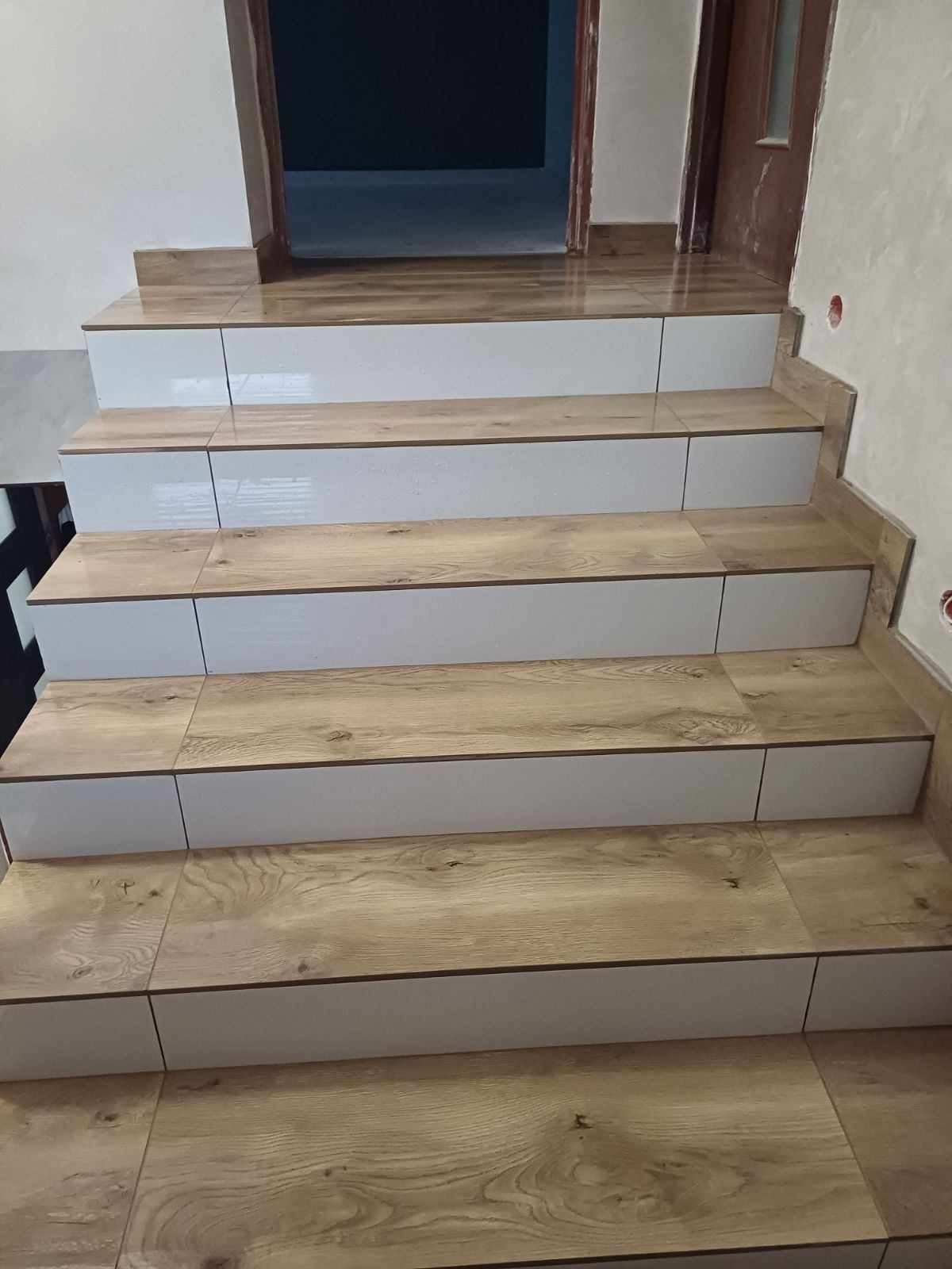 Schody wykończone płytkami imitującymi drewno z białymi panelami bocznymi, prowadzące do drzwi z ościeżnicą w kolorze drewna.