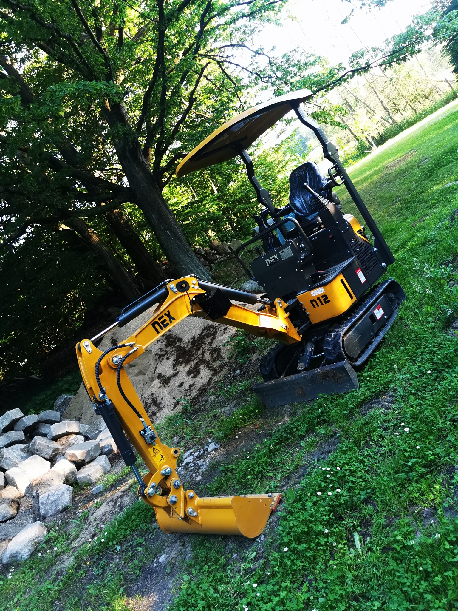 Żółta mini koparka NEX M12 na trawiastym terenie, z wykopanym dołem i stertą kamieni obok, w tle drzewa.