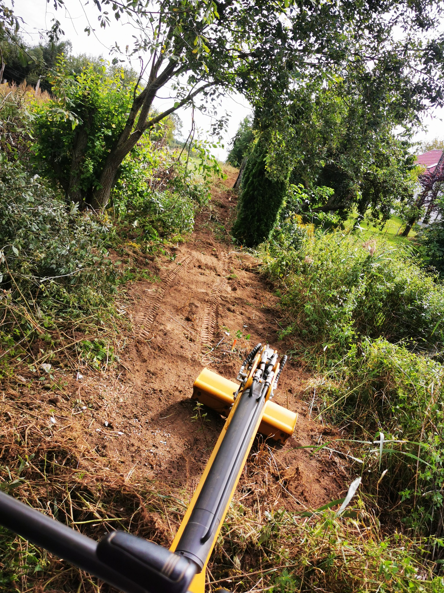 Żółty mini-koparka na zboczu, świeżo wykopana ziemia z widocznymi śladami gąsienic, zarośnięte otoczenie z drzewami i krzewami, widok z perspektywy operatora maszyny.