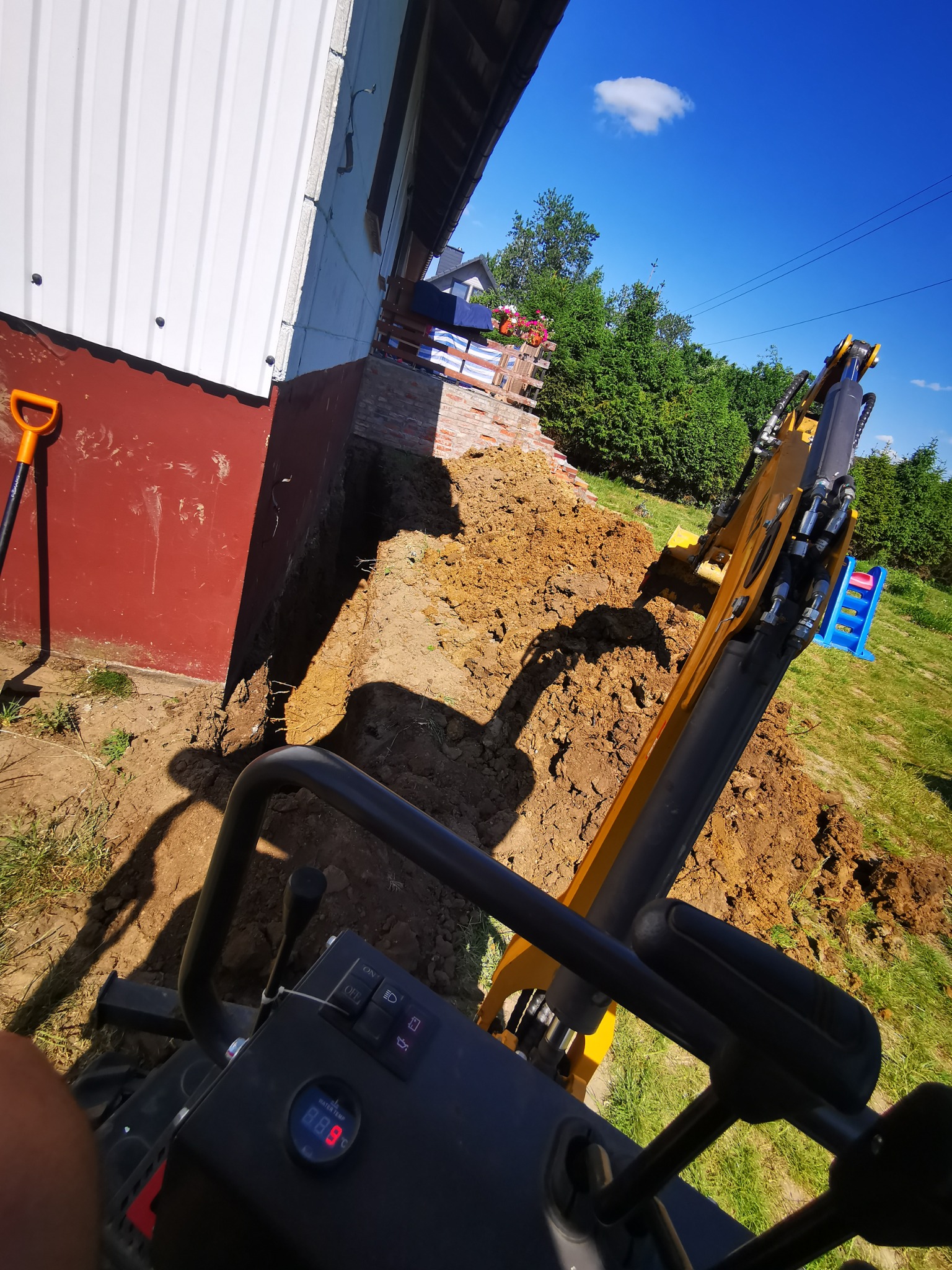Widok z kabiny żółtej mini koparki podczas wykonywania wykopu wzdłuż fundamentu budynku z pomalowaną na czerwono ścianą, na zewnątrz widoczna łopata i fragment placu zabaw.