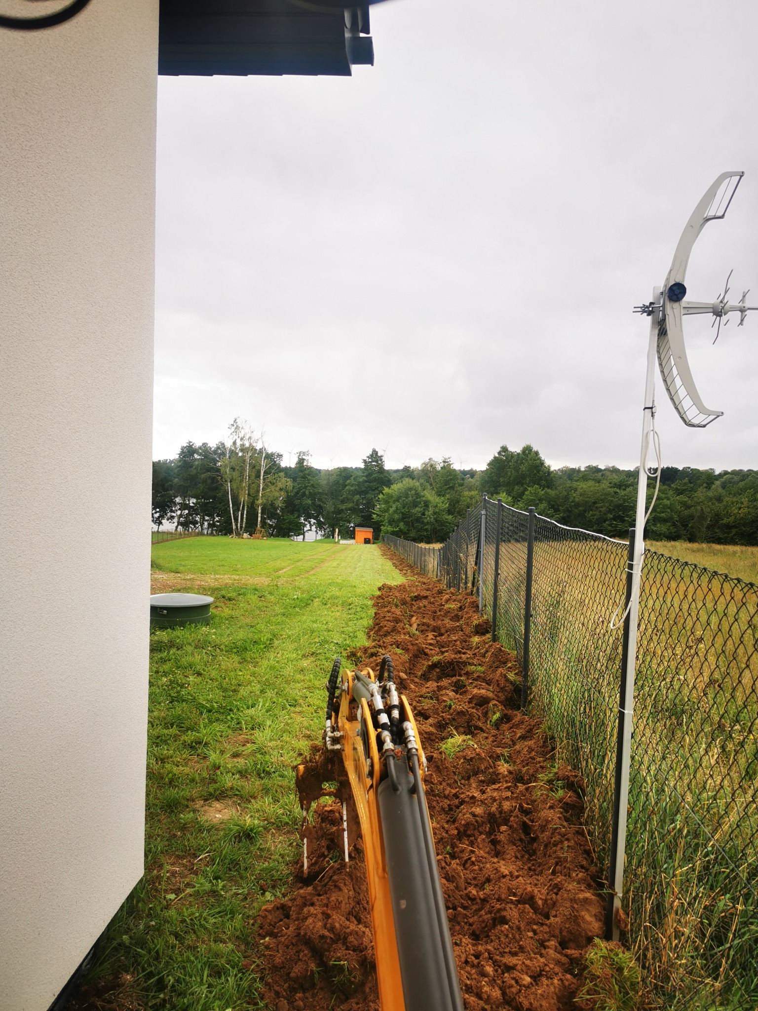 Mini koparka tworzy wykop wzdłuż metalowego ogrodzenia na działce z widokiem na drzewa i antenę satelitarną. Świeżo przekopana ziemia wzdłuż siatki.