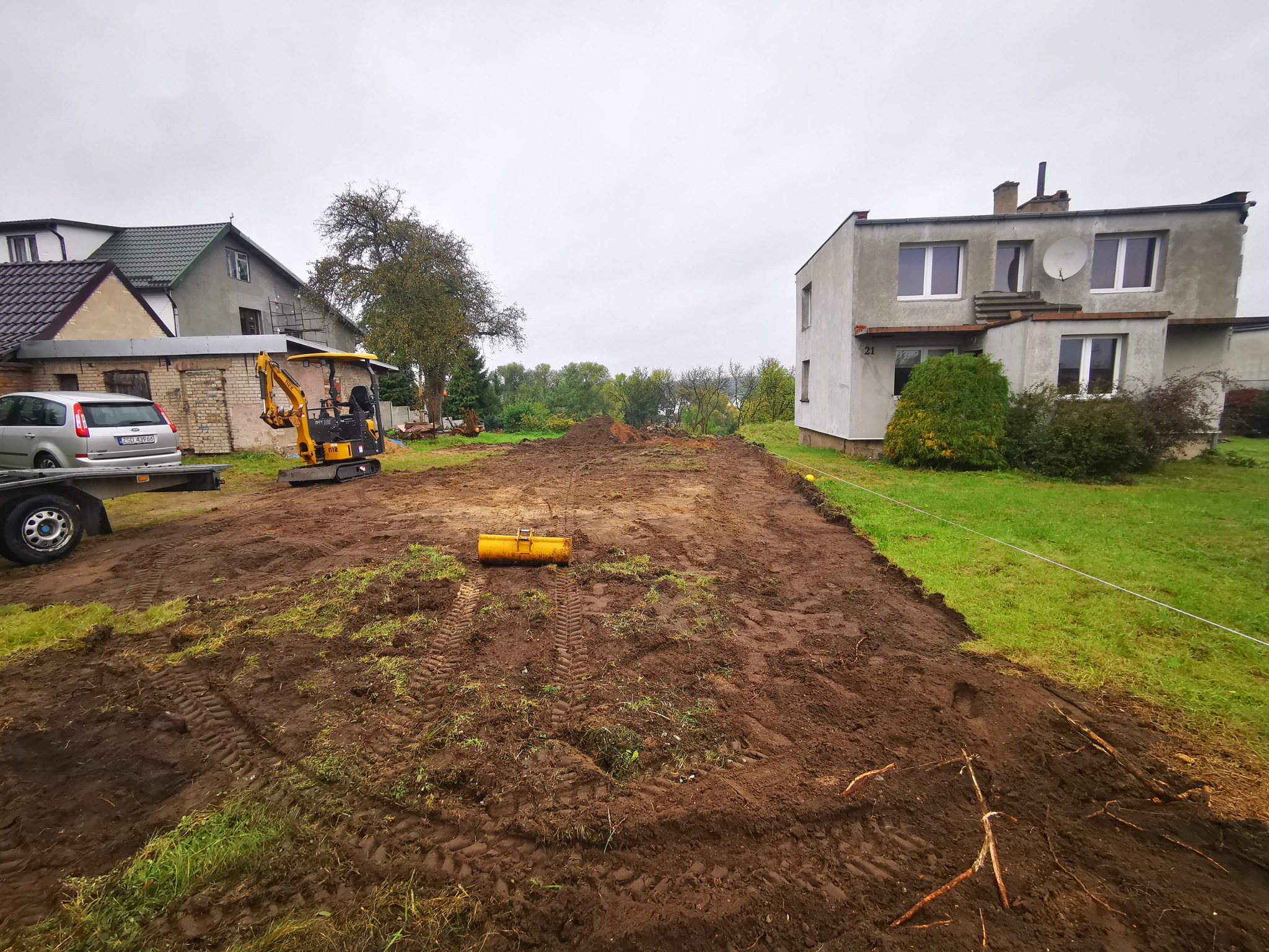 Przygotowany teren budowlany z mini koparką i zagęszczarką w tle, widoczne ślady opon na błotnistej ziemi, domy jednorodzinne w tle, pochmurne niebo.